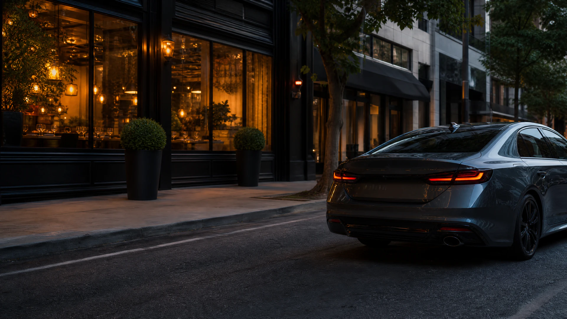A dark grey sedan with glowing tail lights sits on an urban street at dusk in front of a warmly lit restaurant.