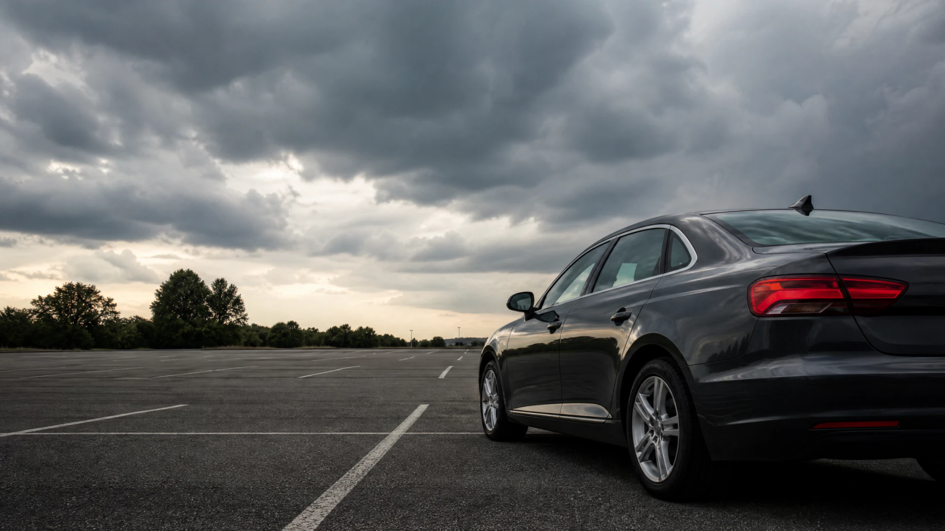A dark grey sedan sits alone in an empty parking lot beneath a stormy, cloud-covered sky.