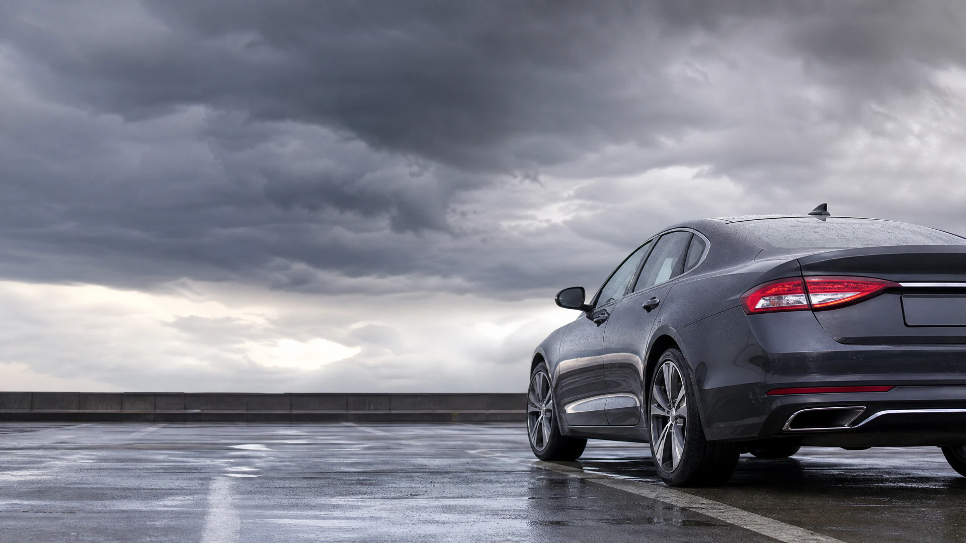 A dark gray sedan sits on a rain-soaked rooftop parking deck beneath a dramatic overcast sky full of storm clouds.