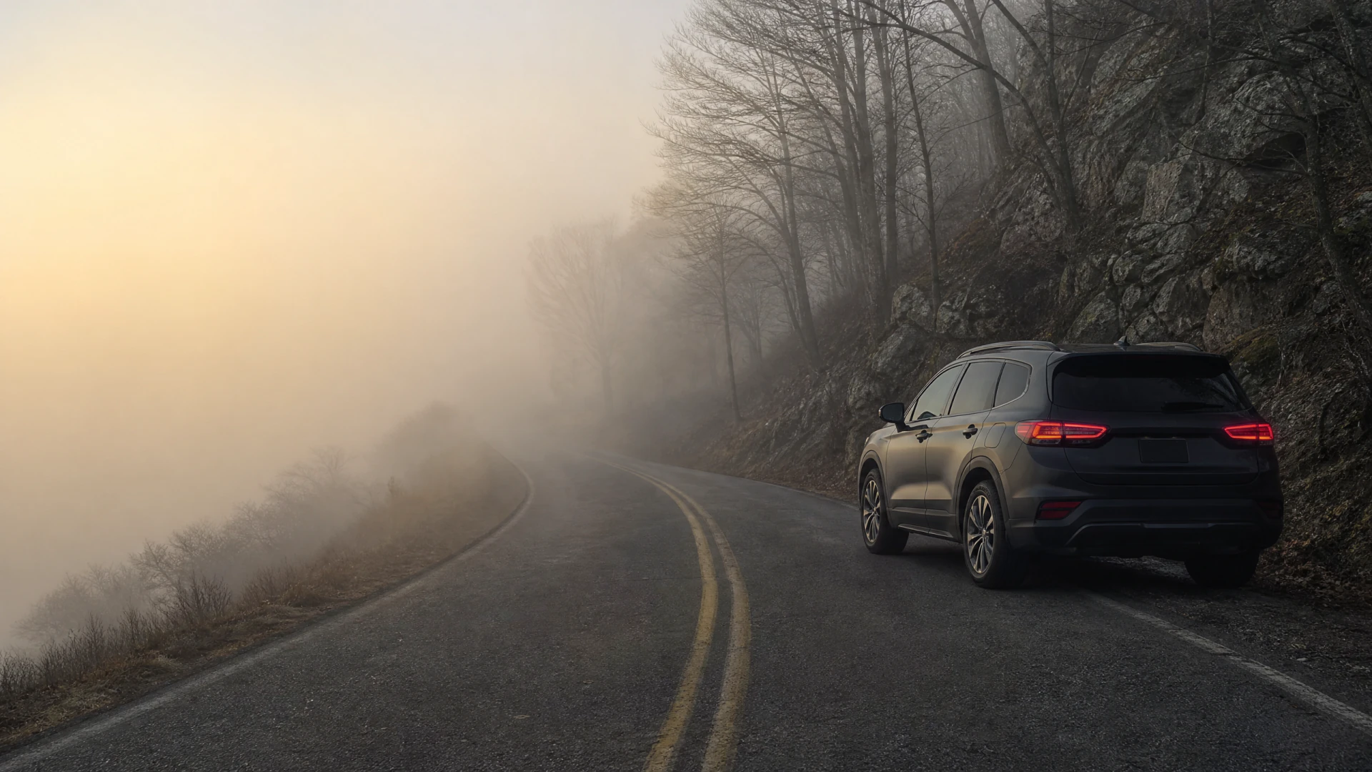 A dark SUV with glowing tail lights sits on the edge of a foggy, winding mountain road flanked by bare trees and rocky outcroppings.