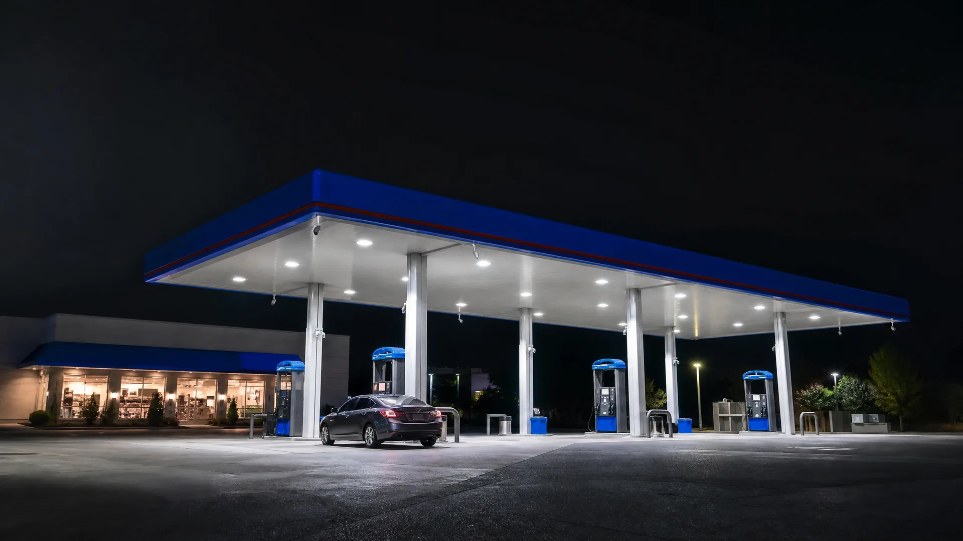 A lit gas station canopy with blue fascia and white underside at night, with fuel pumps, a dark sedan parked beneath it, and a convenience store with lit windows at left.