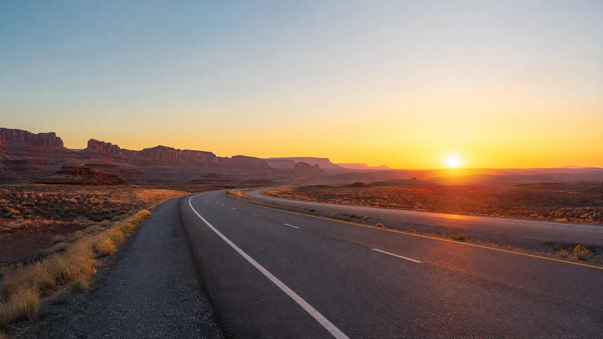 A two-lane paved highway curves through flat desert scrub toward distant mesas, with the sun low on the horizon at dawn or dusk.