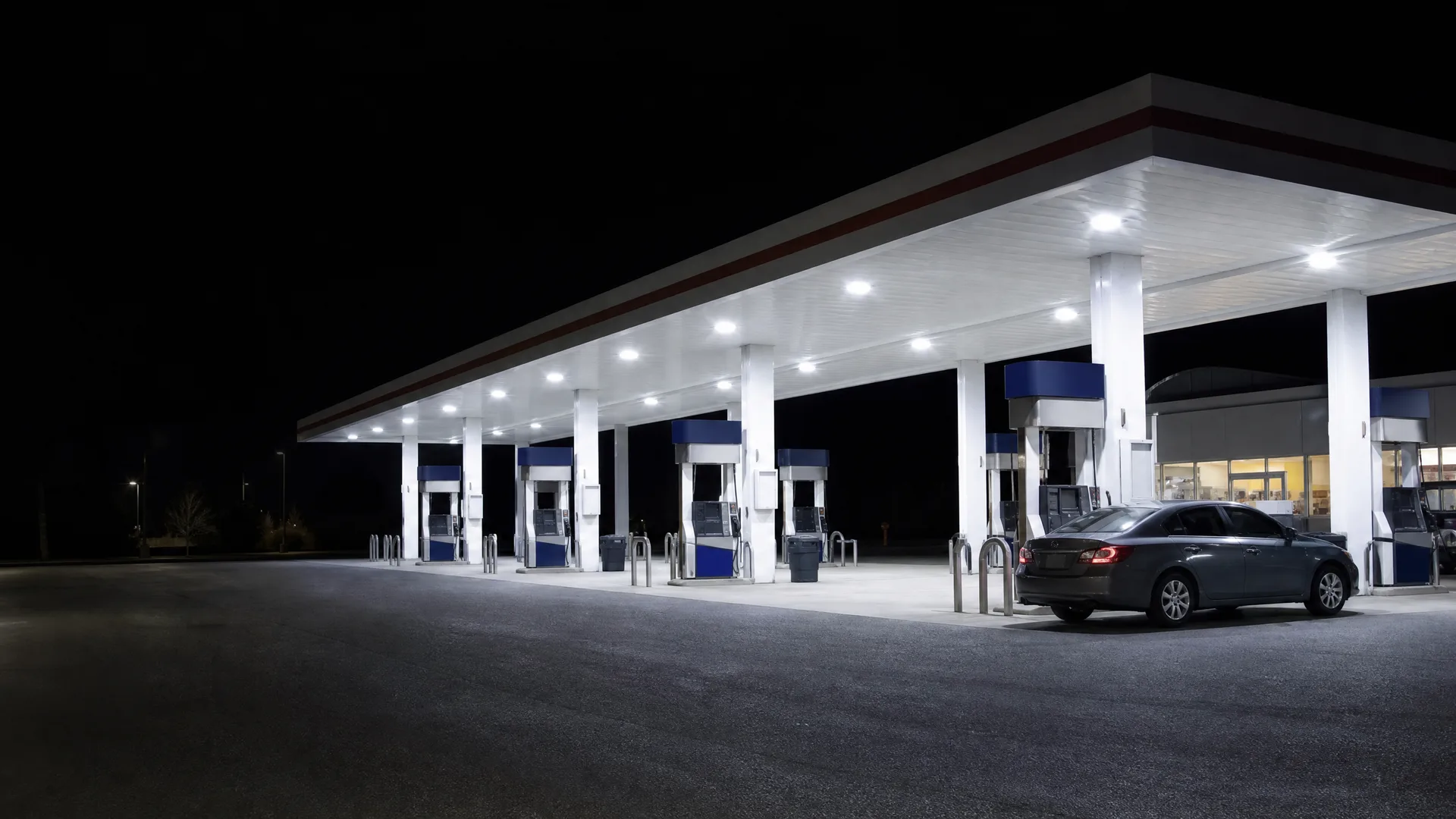 A brightly lit gas station canopy with a row of blue and white fuel pumps at night, with a single dark sedan parked at the far end and empty asphalt in the foreground.
