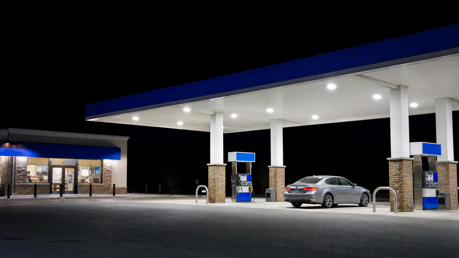 A lit gas station canopy with blue trim at night, with a silver sedan parked beside a fuel pump and a convenience store at left.