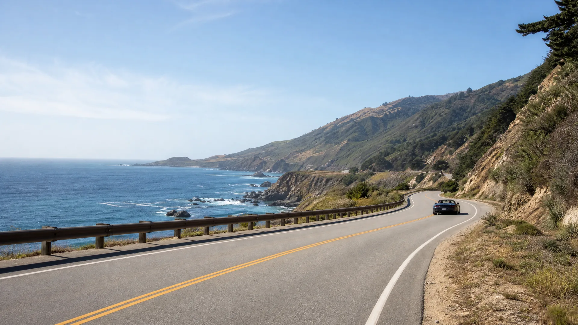 A two-lane coastal highway with a guardrail curves along a hillside above the ocean, with a dark car driving away in the distance and brush-covered slopes on the right.