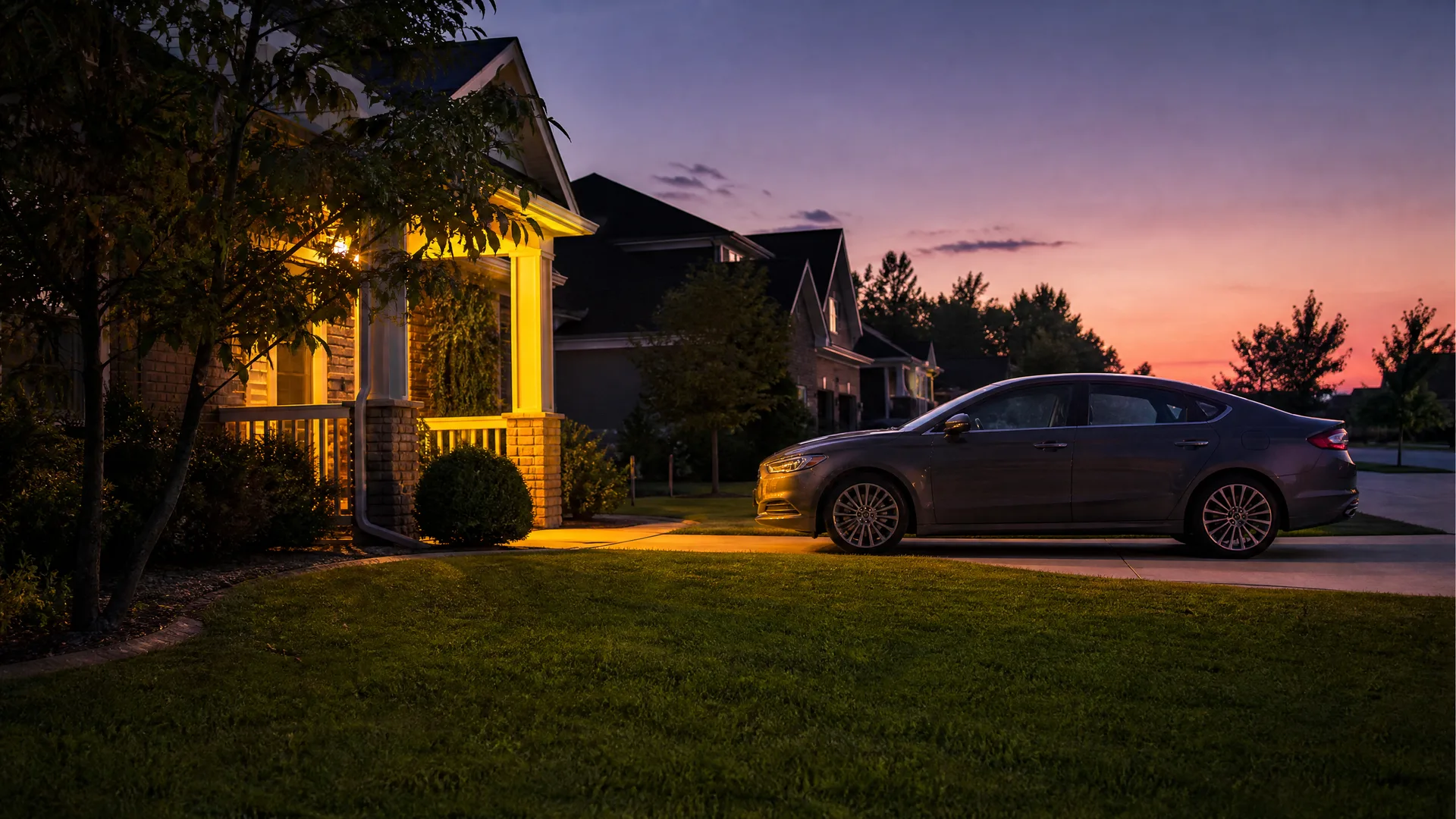 A dark gray four-door sedan parked on a driveway beside a brick house with a lit porch at dusk, with a mowed lawn in the foreground and an orange and purple sky.