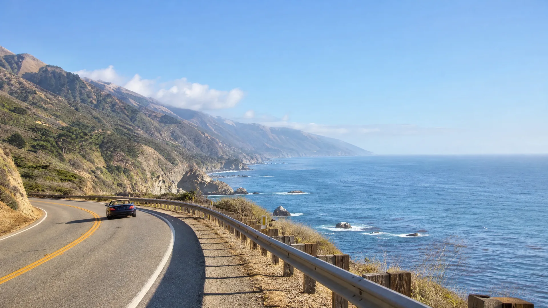A dark convertible car drives on a curving two-lane coastal highway with a wooden guardrail, beside a blue ocean and steep hills under a clear sky.