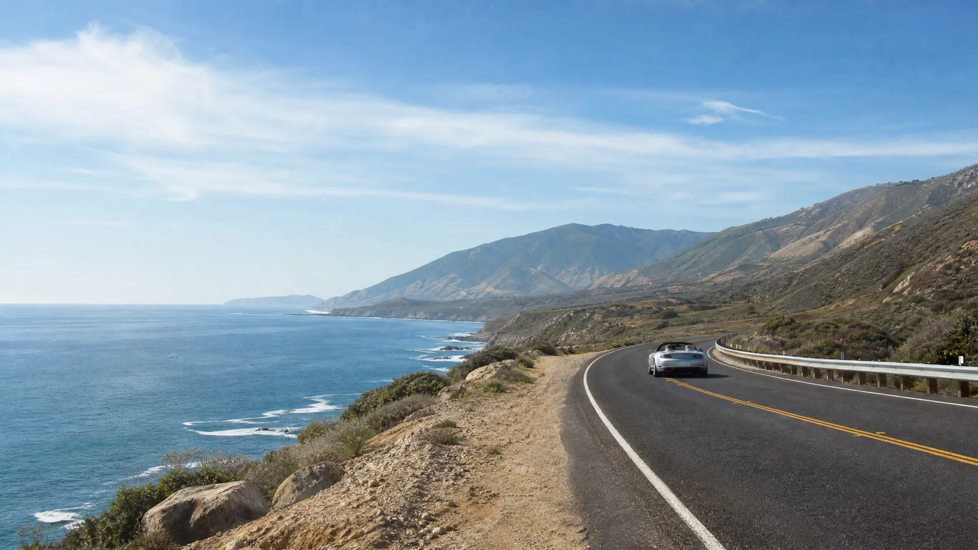 A convertible car drives along a two-lane coastal highway with a guardrail, with the ocean and dry hillsides to the left and mountains ahead under a mostly clear sky.