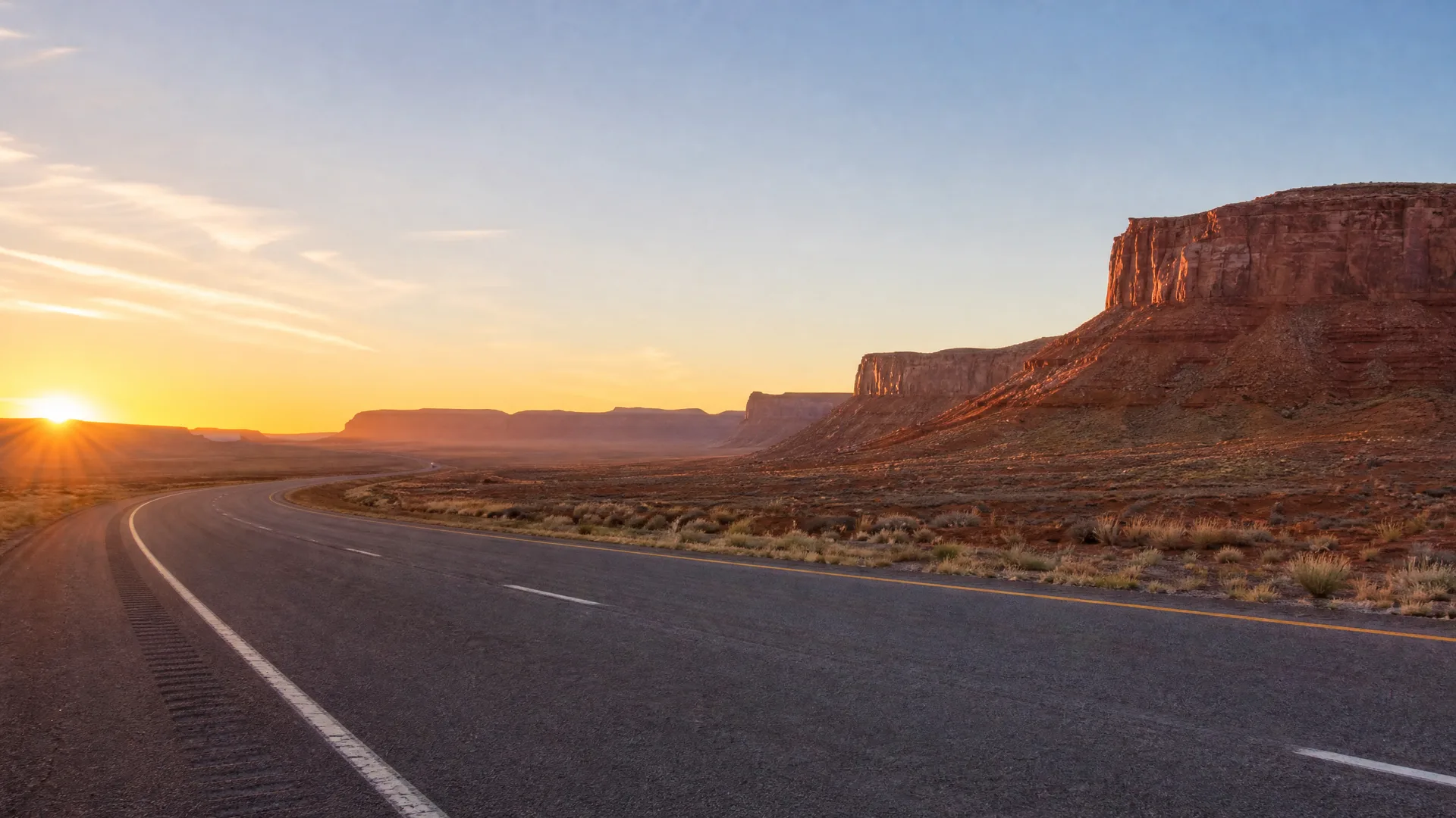 A two-lane asphalt road with white and yellow markings curves through desert scrub toward a flat-topped red rock mesa, with the sun low on the horizon at left.