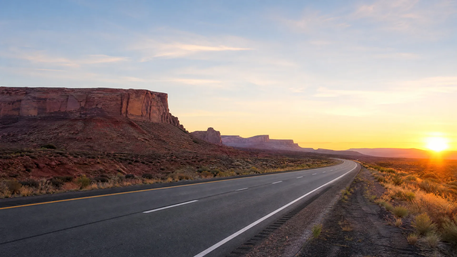 A two-lane paved highway curves through desert scrubland toward the setting sun, with a flat-topped red rock mesa and distant cliffs on the left.