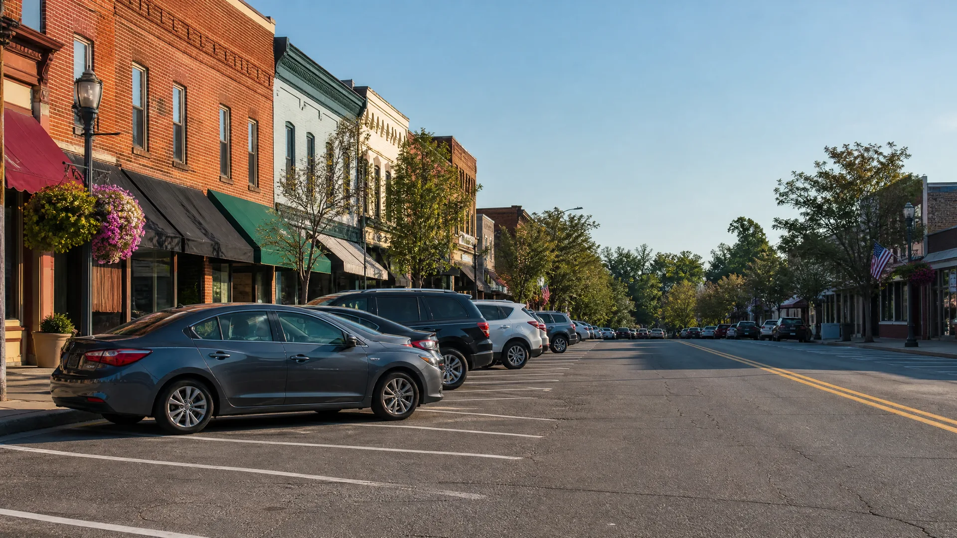 A small-town main street with angled parking, cars along a curb beside two- and three-story brick commercial buildings with awnings, trees lining the far side, and a US flag on a building at right.