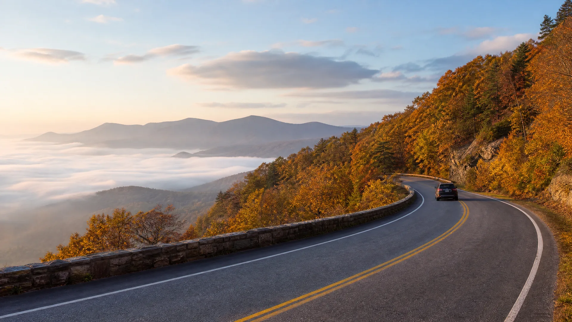 A curving two-lane mountain road with a stone guardrail and a single dark SUV, bordered by autumn-colored trees, overlooking fog-filled valleys and distant ridges.