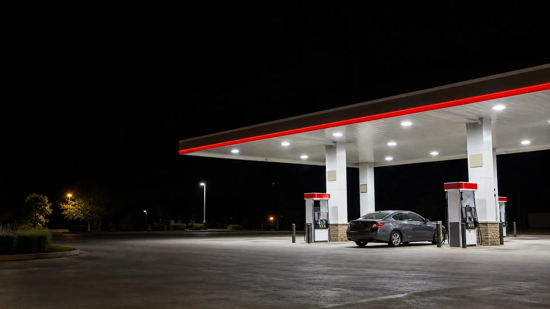 A lit gas station canopy with red trim at night, with one gray sedan parked beside a fuel pump on an empty concrete lot.