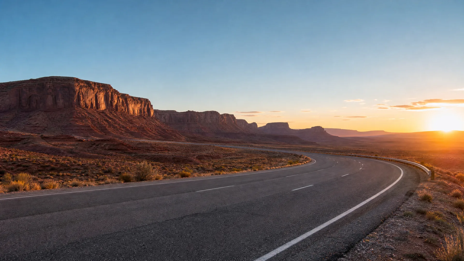 A two-lane paved road curves to the right through open desert scrubland, with layered mesas and cliffs on the horizon and the sun low in a clear sky.