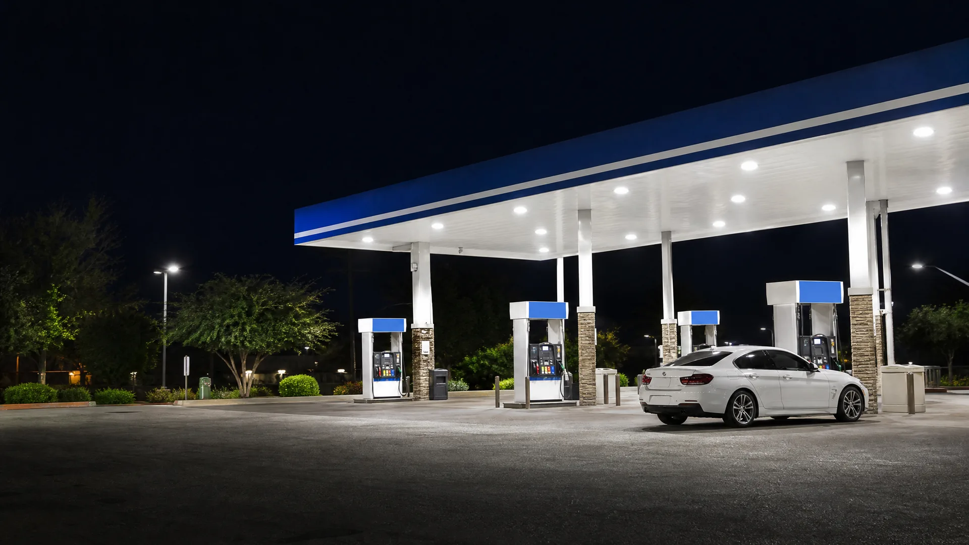 A white sedan parked at a lit gas station canopy with blue-topped fuel pumps at night, with trees and a dark sky behind.