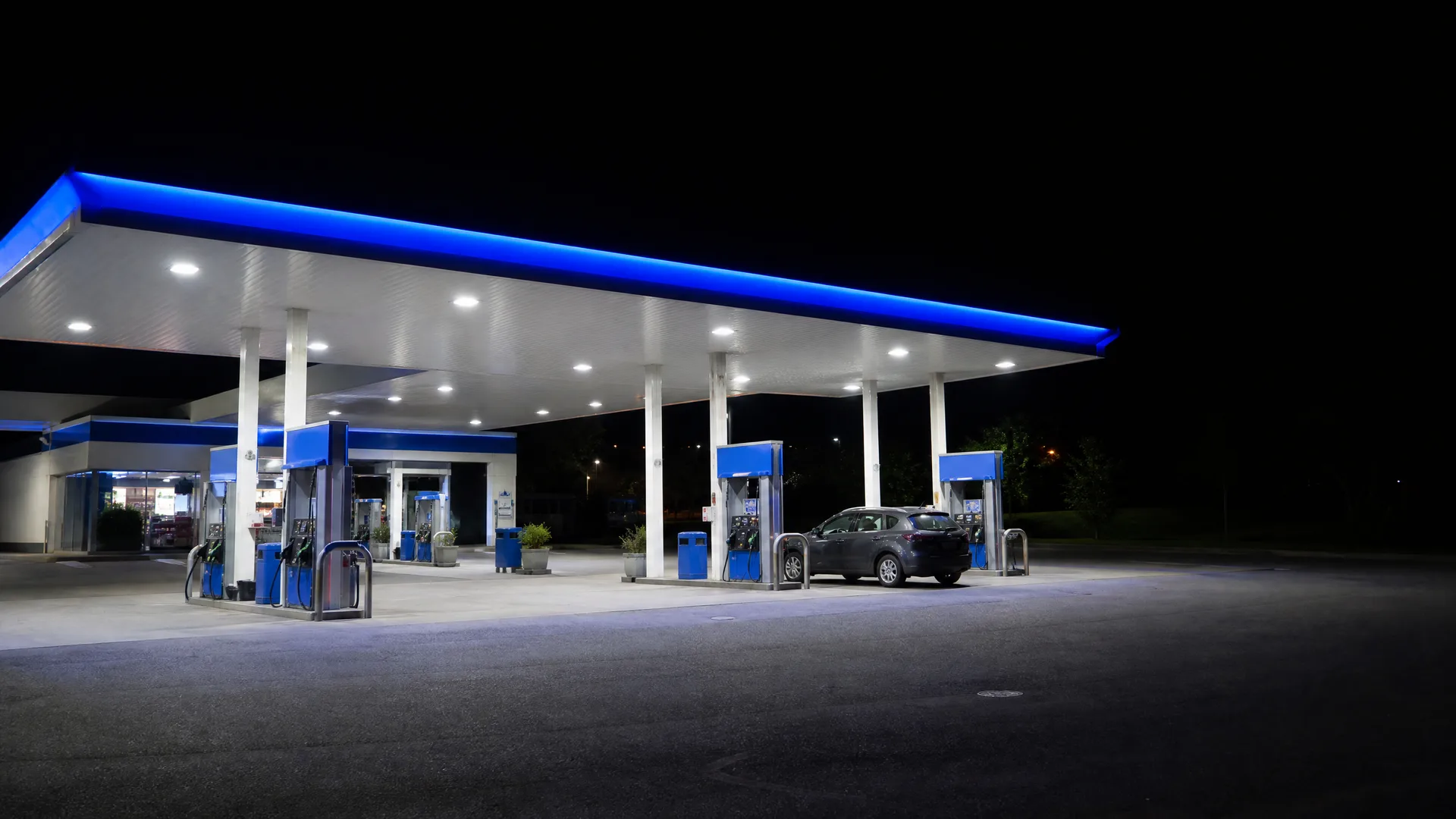 A gas station at night with a blue-trimmed, brightly lit canopy over several fuel pumps, an SUV parked at one pump, and an empty paved forecourt in the foreground.