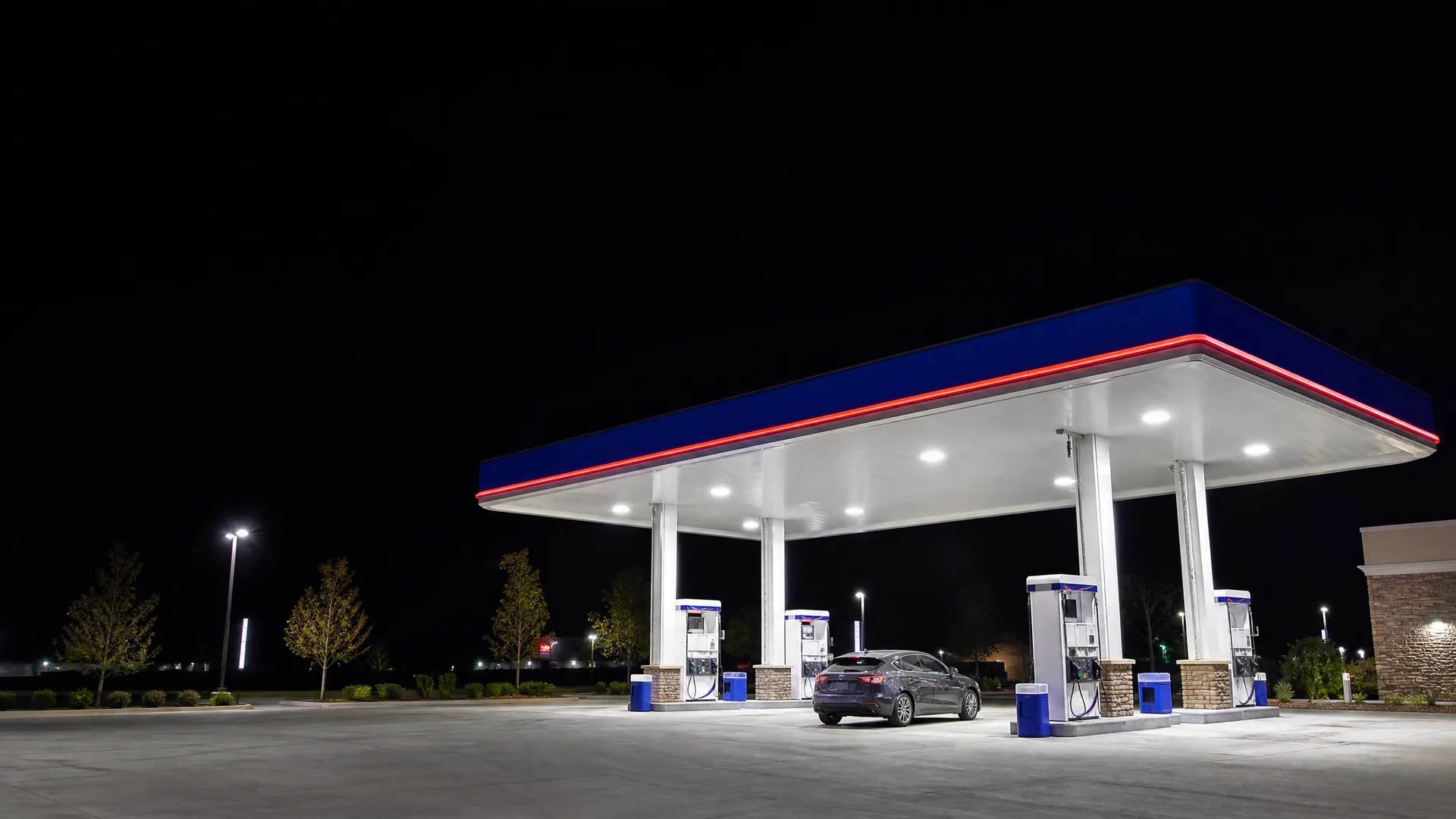 A lit gas station canopy with blue fascia and red trim at night, with fuel pumps and a dark sedan parked beneath it.