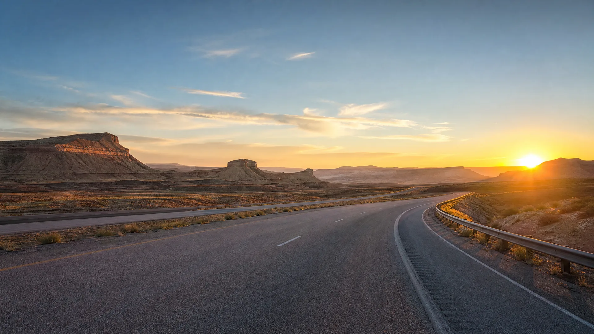 A two-lane paved highway curves through arid scrubland with flat-topped mesas in the distance as the sun sets near the horizon.