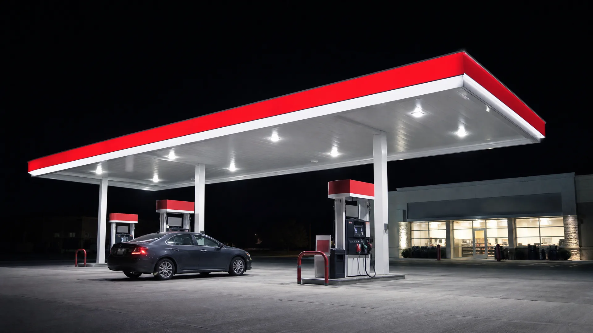 A gas station at night with a red and white lit canopy, fuel pumps, a dark sedan parked beneath it, and a convenience store with lit windows to the right.