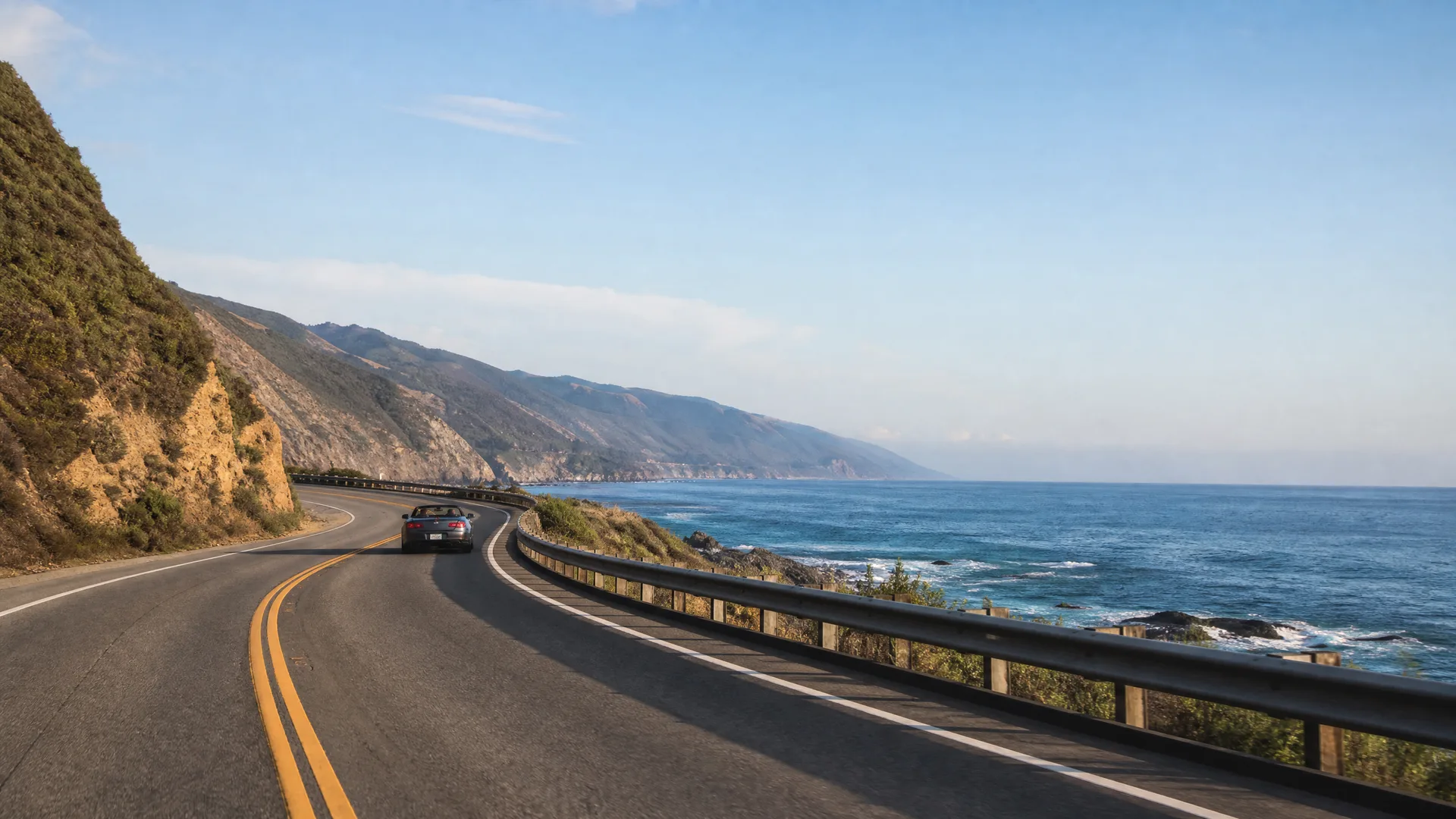 A dark convertible car drives along a curving two-lane coastal highway with a metal guardrail, with cliffs on the left and the ocean and headlands on the right.