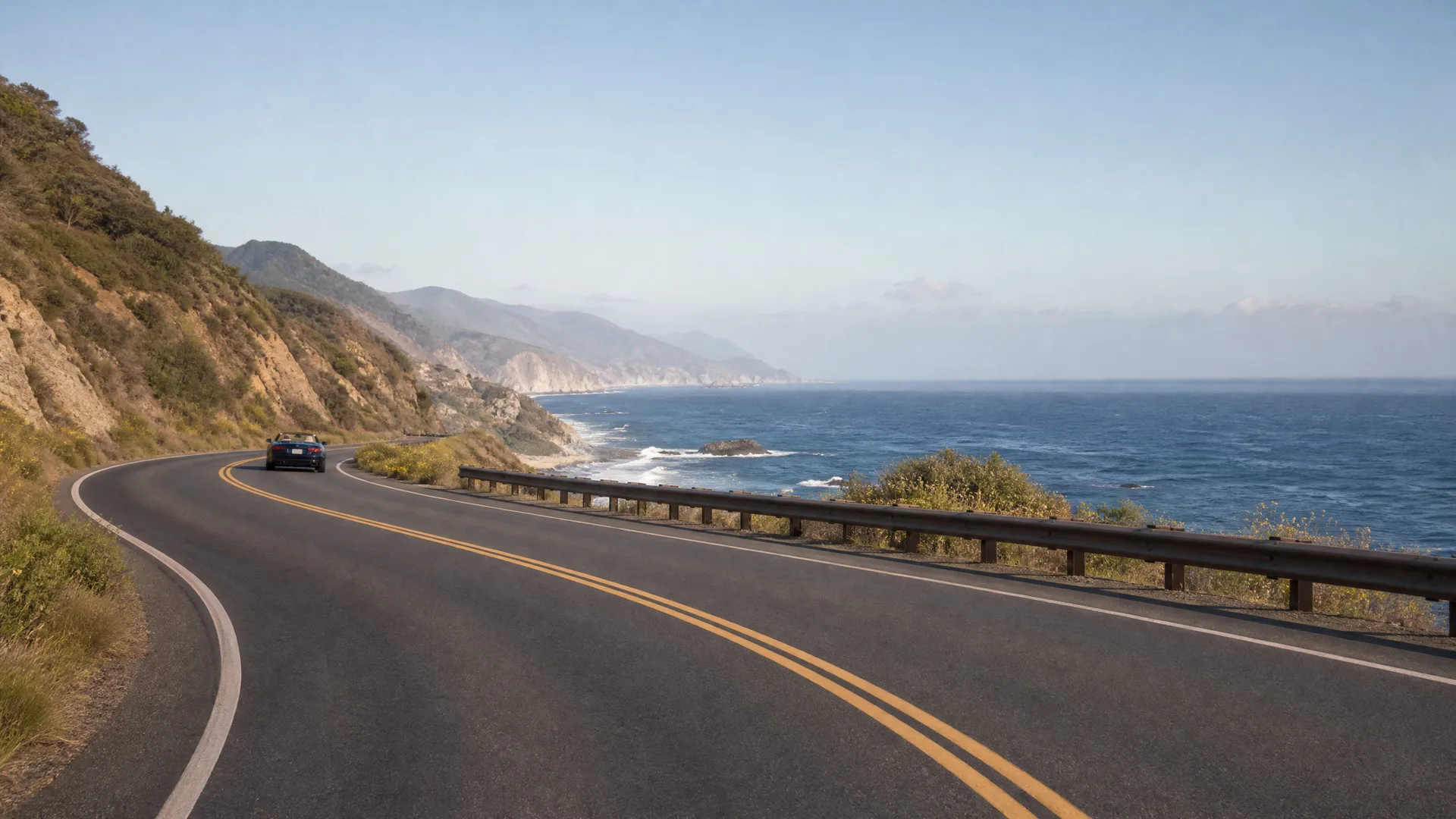 A two-lane coastal highway with double yellow center lines curves along a hillside above the ocean, with a dark car driving away in the distance and a metal guardrail on the seaward side.