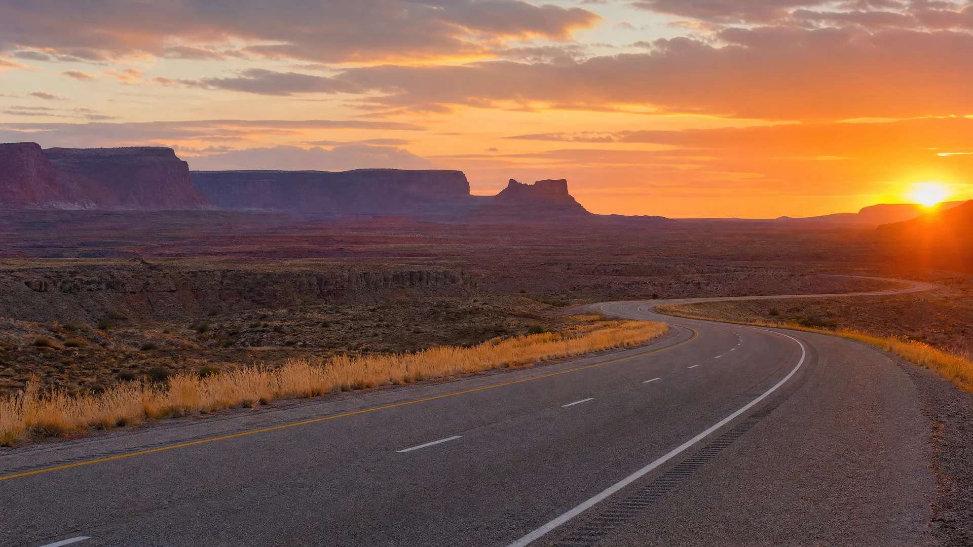A two-lane paved highway curves through flat desert scrub toward distant mesas and buttes under an orange sky at sunset.