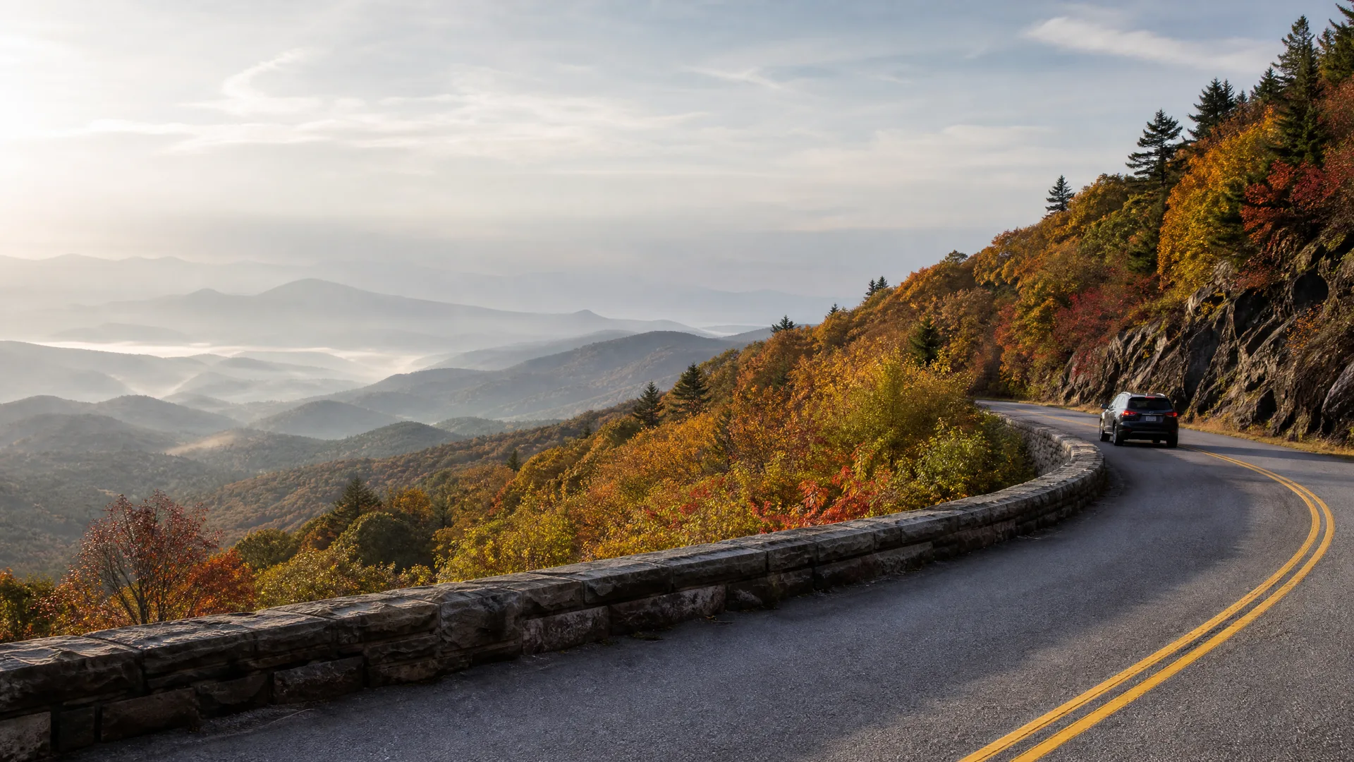 A curving two-lane mountain road with a double yellow center line and a low stone wall, bordered by autumn-colored trees, with a dark SUV driving ahead and layered hazy ridges in the distance.
