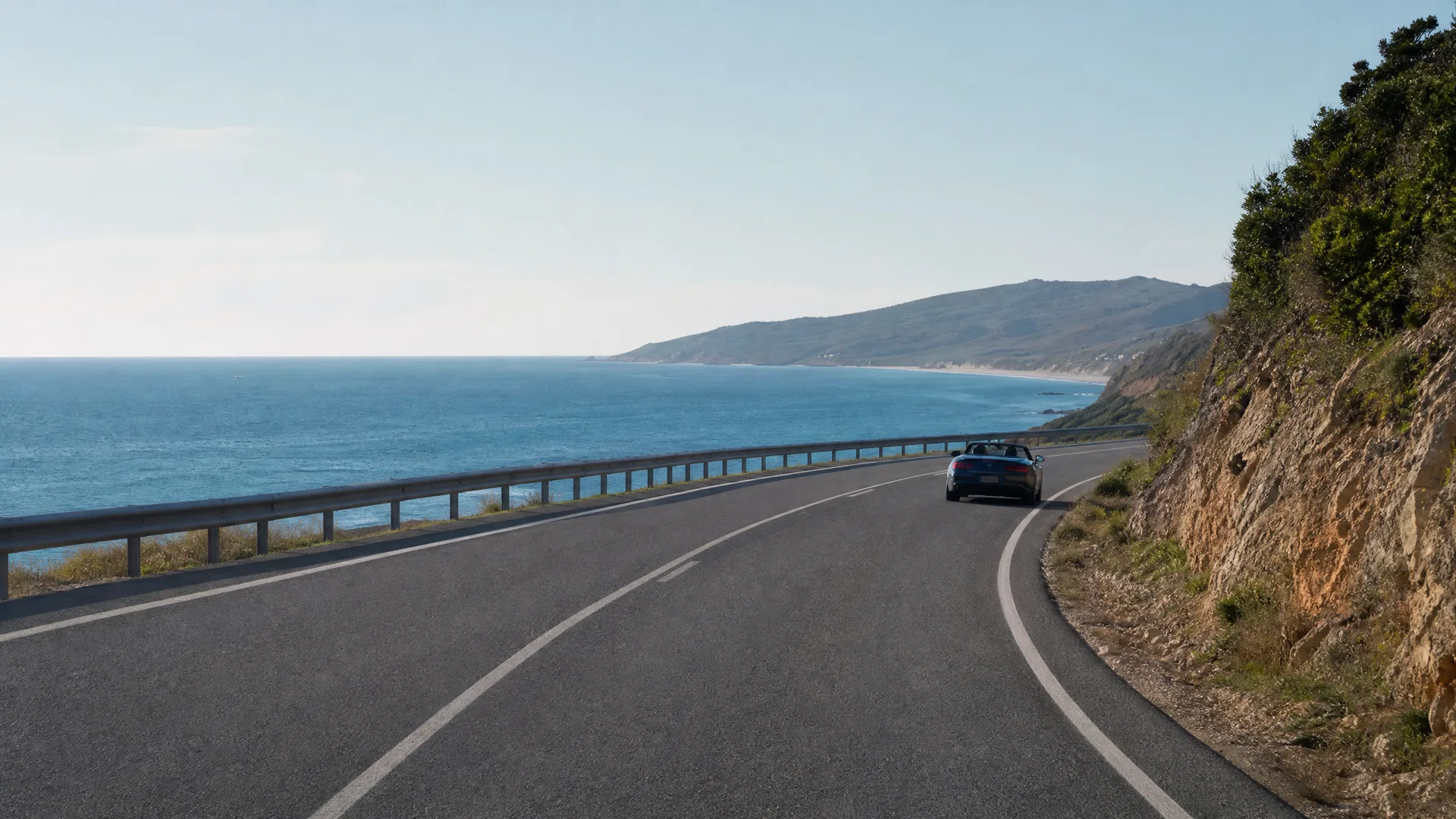 A dark convertible car drives along a curving two-lane coastal road with a guardrail on the left above the sea and a rocky vegetated slope on the right.