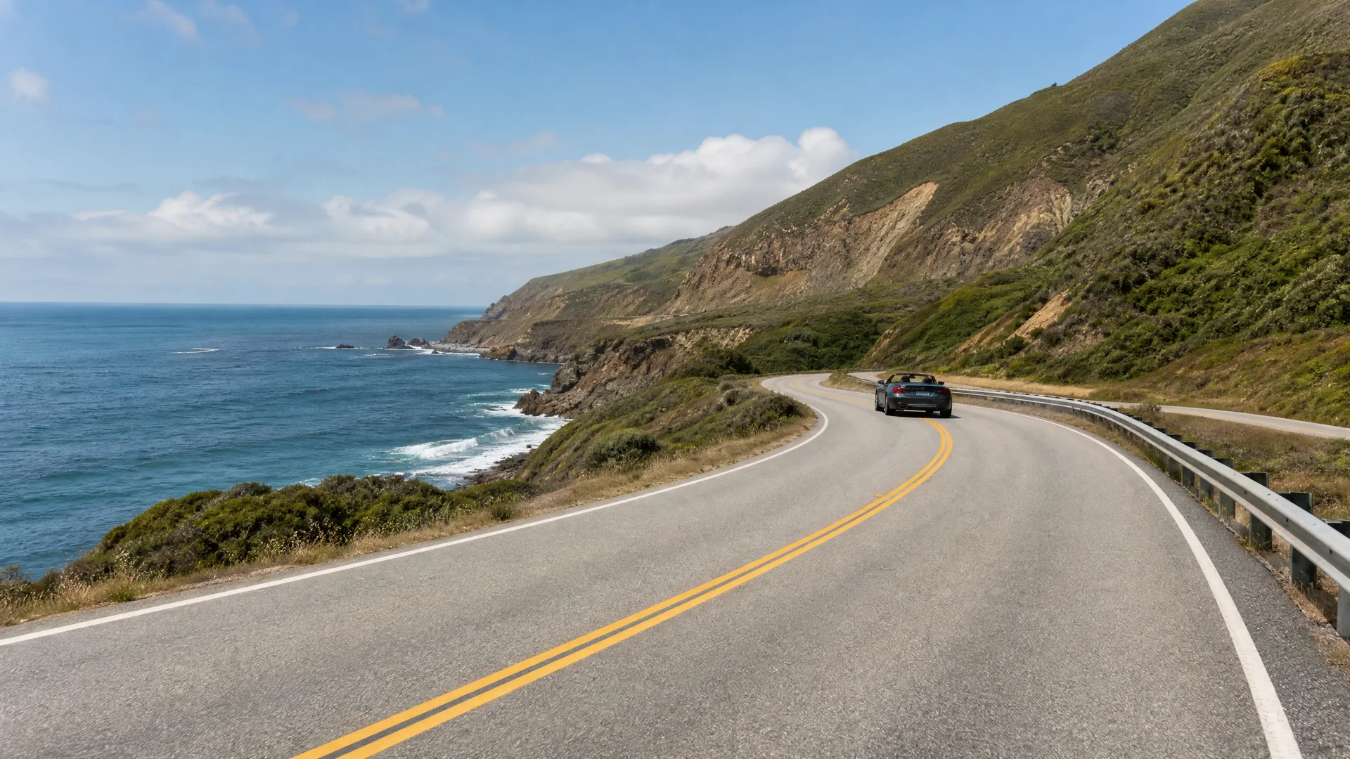 A dark convertible car drives along a curving two-lane coastal highway with double yellow center lines, the ocean and cliffs on the left and steep green hillsides with a guardrail on the right.