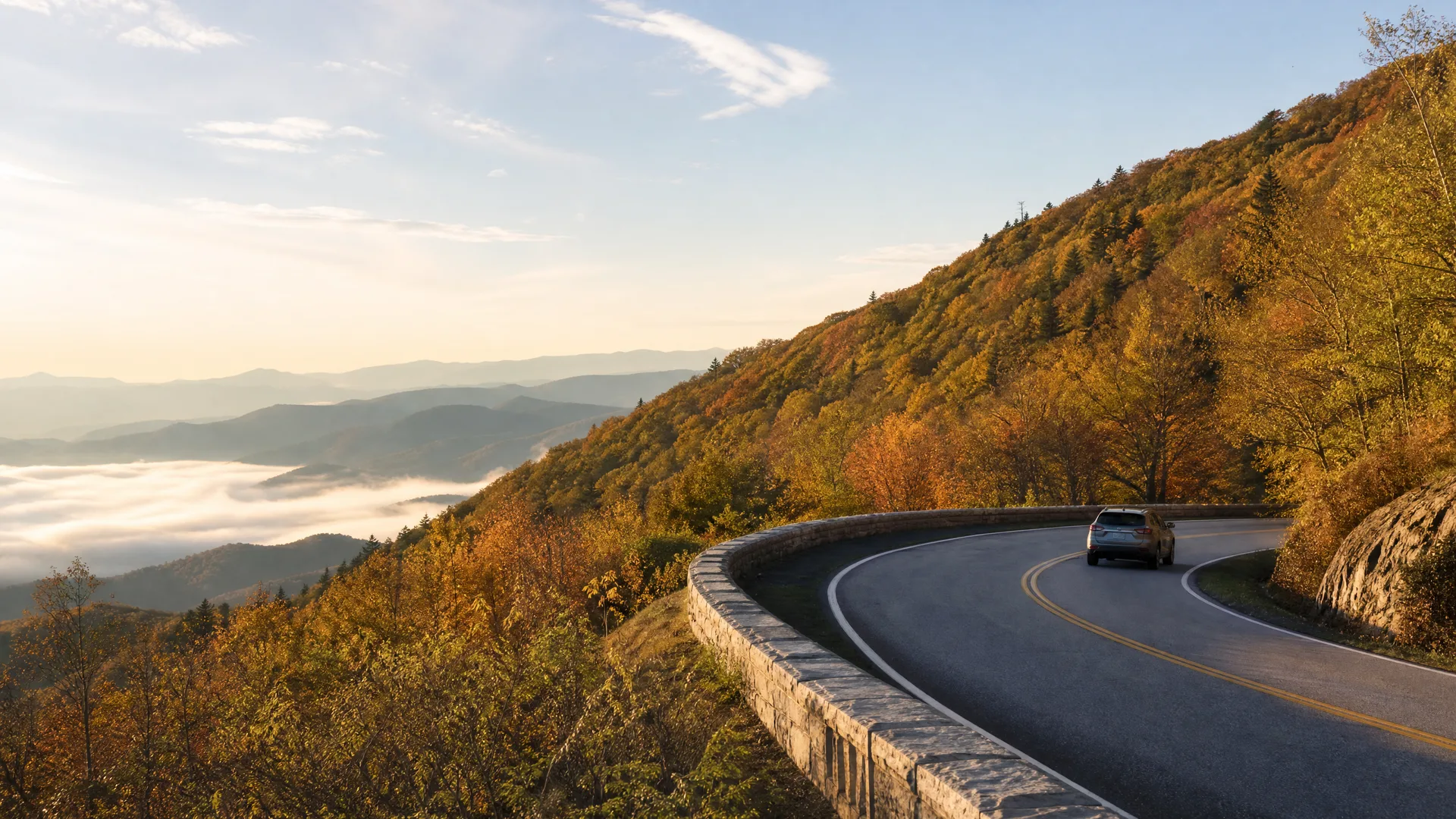 A car drives on a curving two-lane mountain road with a low stone wall, bordered by autumn-colored trees, with fog-filled valleys and ridgelines visible in the distance.