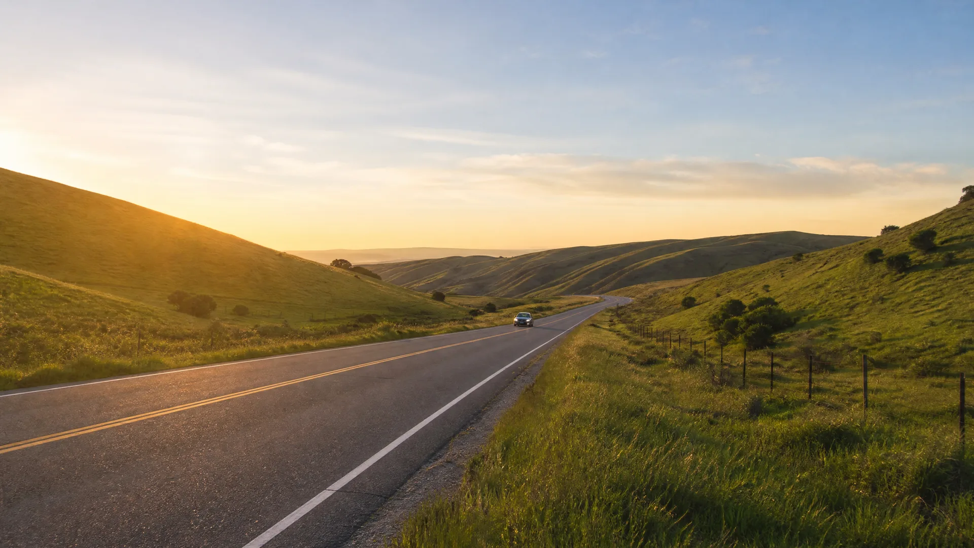 A two-lane road with yellow center lines curves through green rolling hills at sunrise, with a single light-colored car in the distance and a wire fence along the right shoulder.