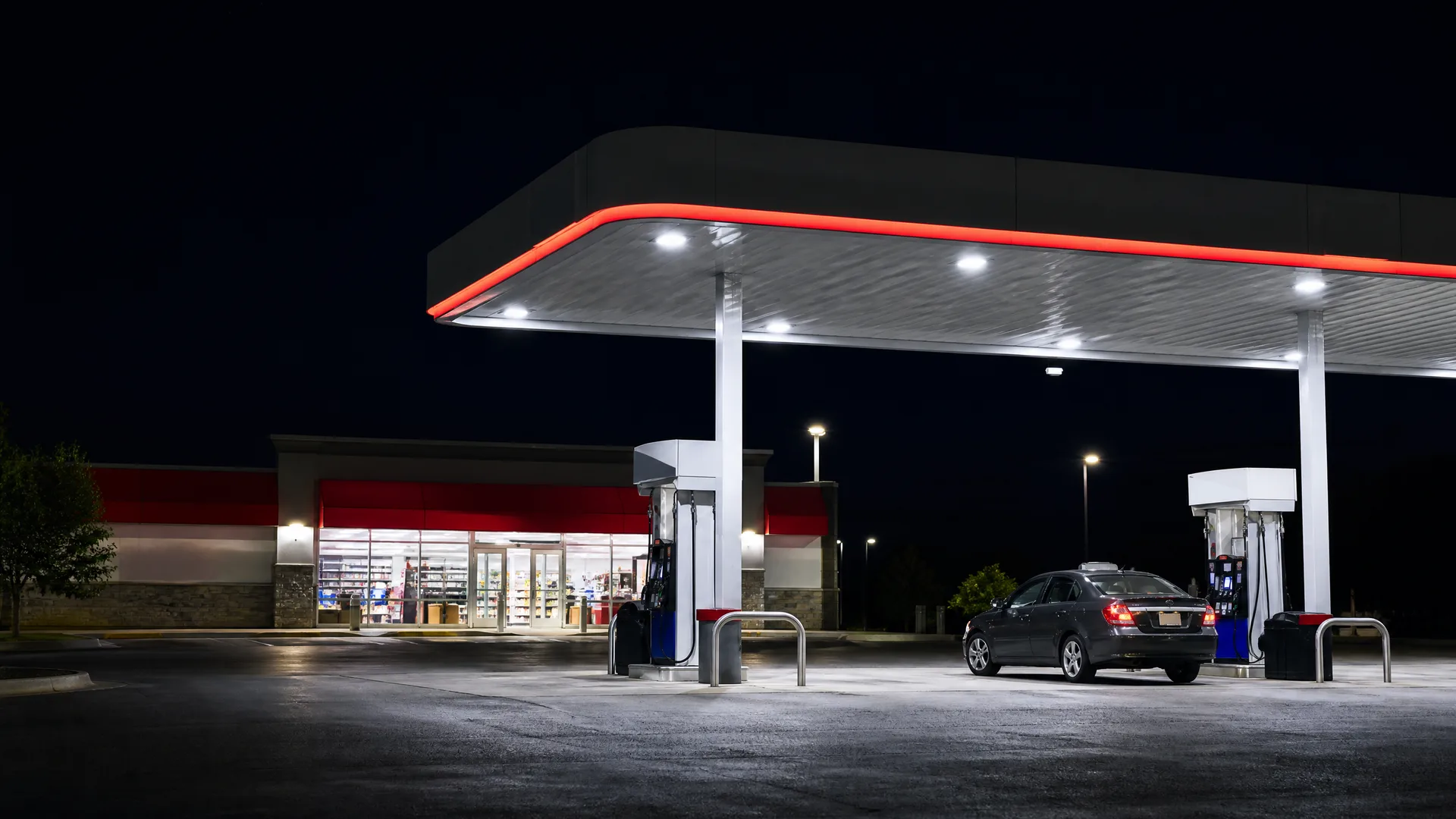 A lit gas station canopy with two fuel pumps at night, a dark sedan parked at the right pump, and a convenience store with red trim in the background.