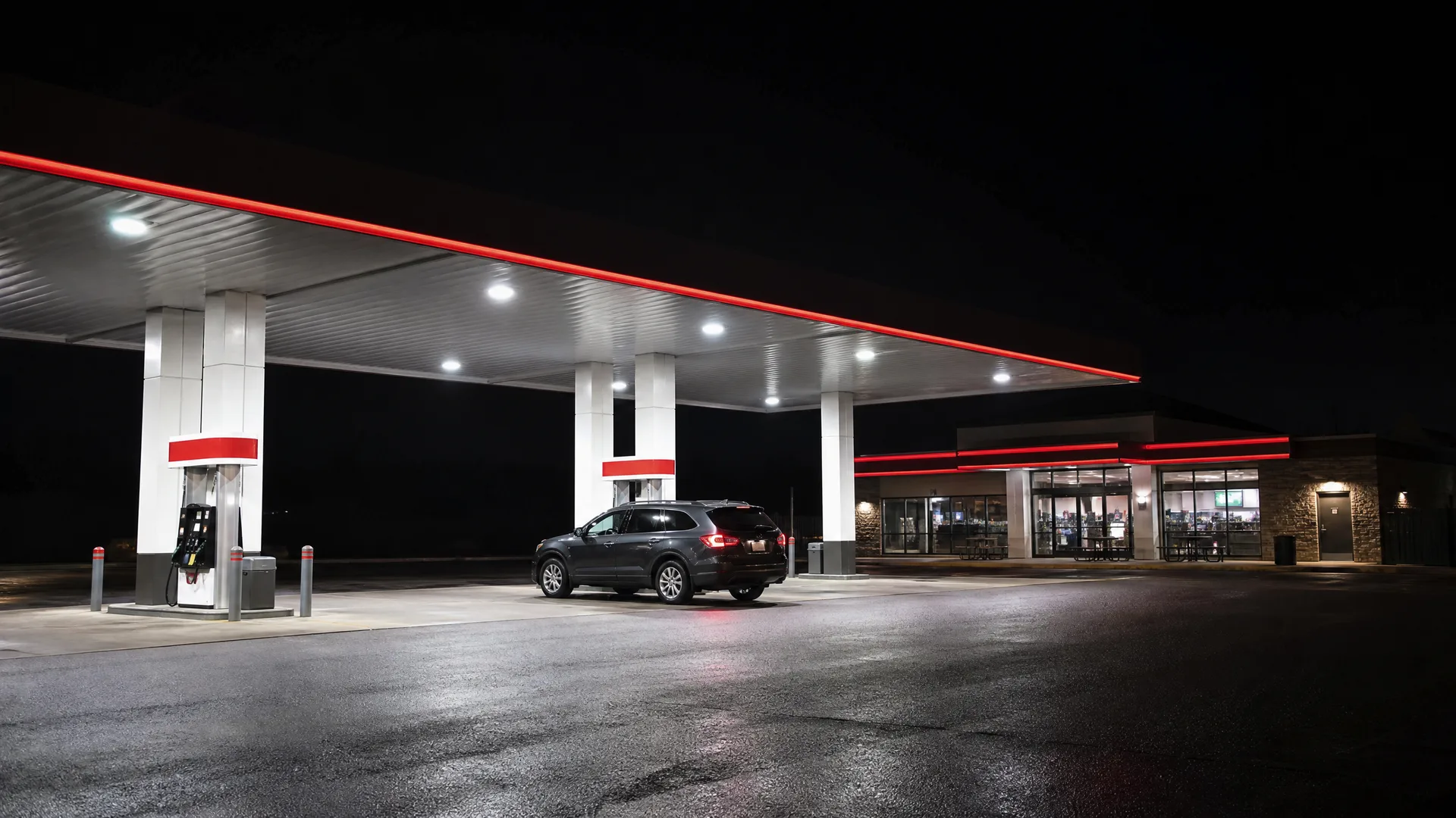 A gas station at night with a lit canopy edged in red, a dark SUV parked near the pumps, and a convenience store with lighted windows in the background.