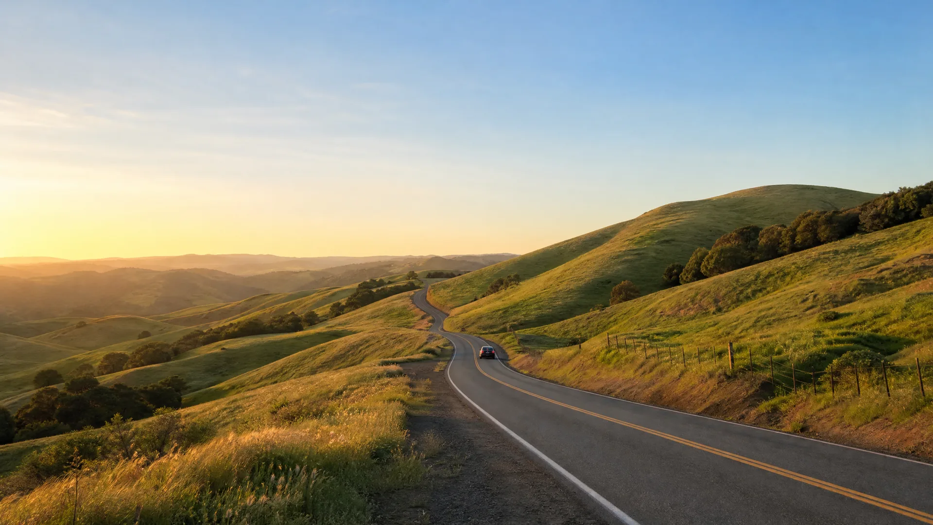 A two-lane road with a double yellow center line curves through rolling green and golden hills at low sun, with a single car in the distance and a wire fence along the right shoulder.