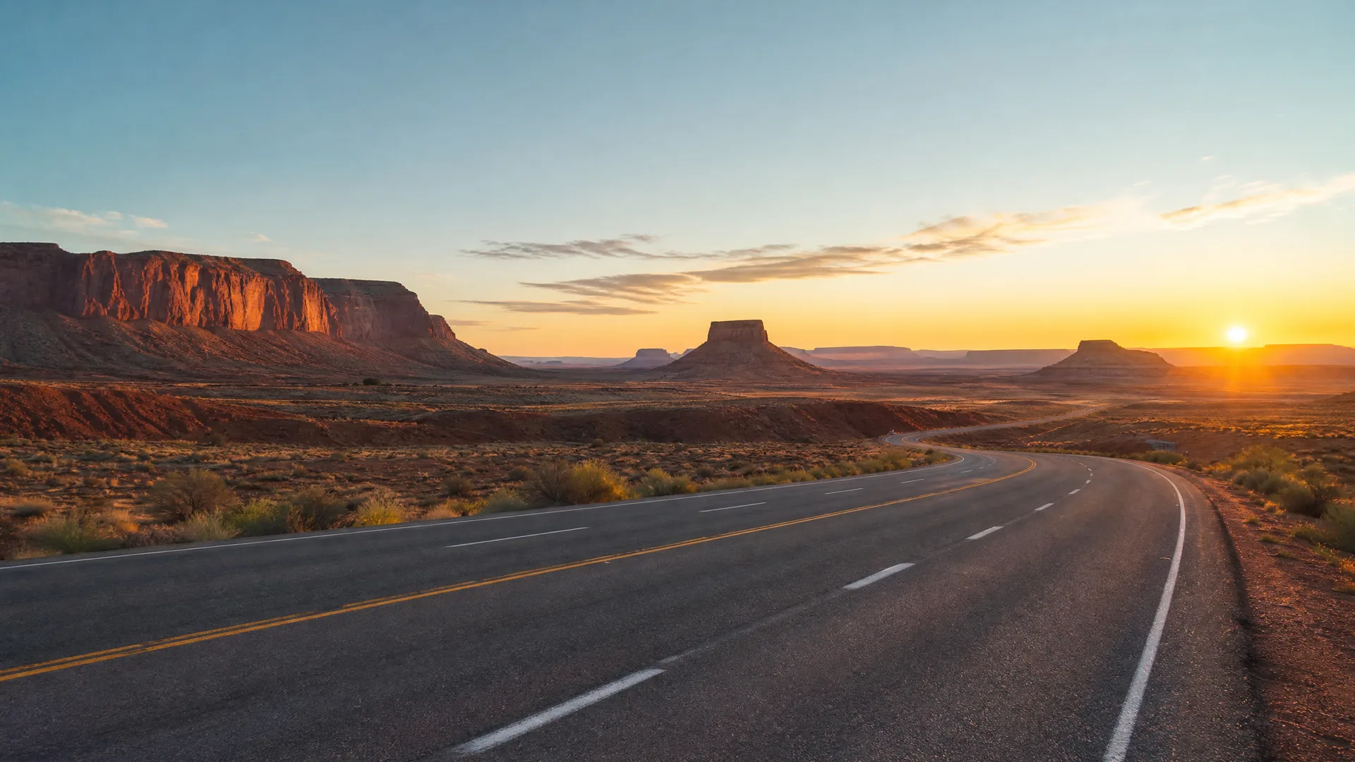 A two-lane highway with yellow center lines curves to the right through desert scrub toward mesas and buttes, with the sun low on the horizon.