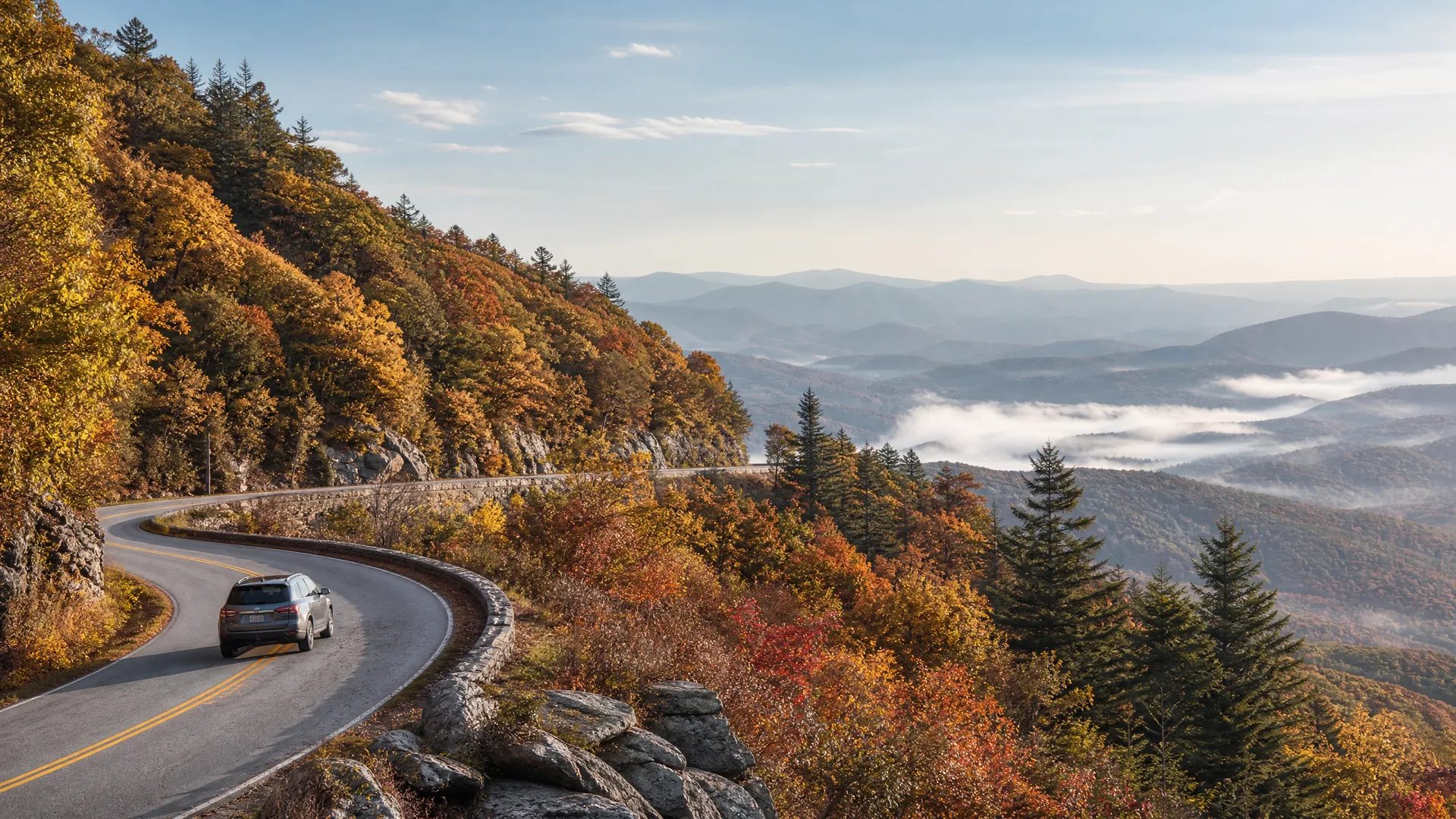 A dark SUV drives on a curving two-lane mountain road with a stone wall, bordered by trees in autumn colors, with fog-filled valleys and ridgelines in the distance.