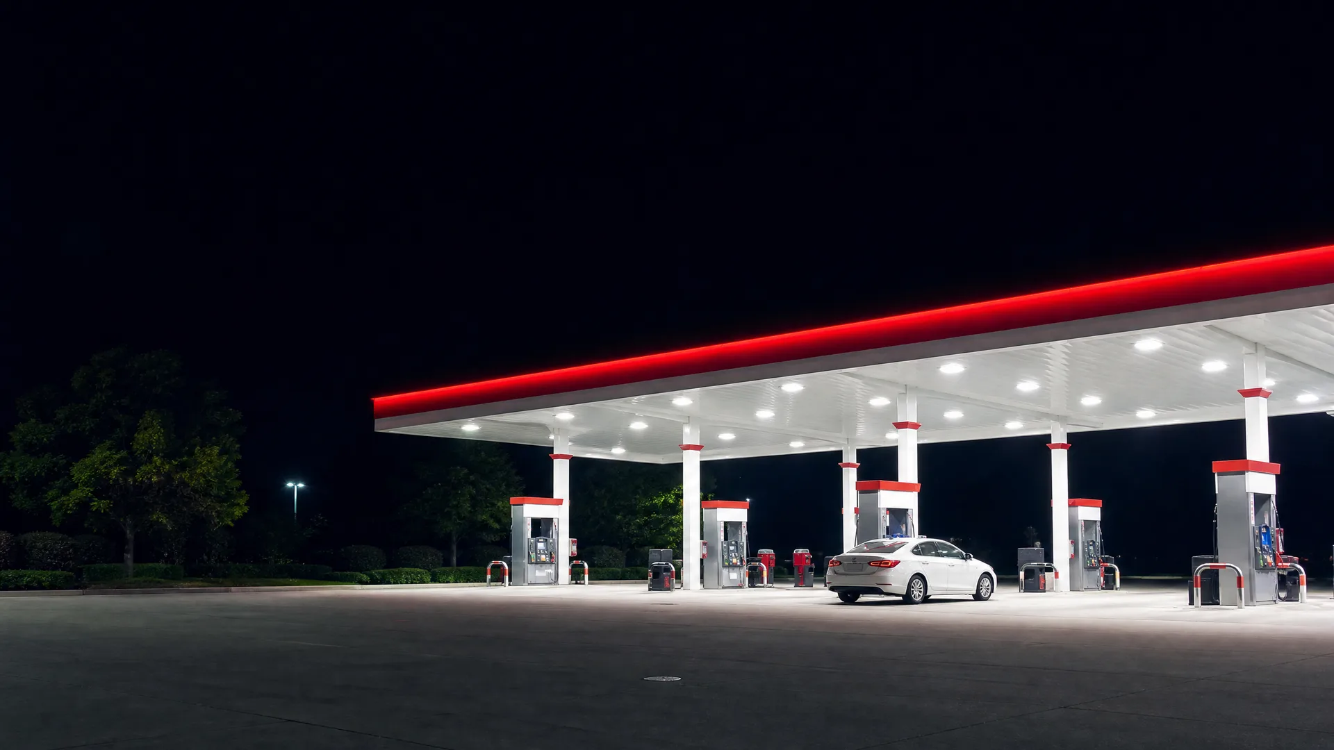 A lit gas station canopy with a red edge and white columns at night, with a white sedan parked beside one of the fuel pumps on empty pavement.