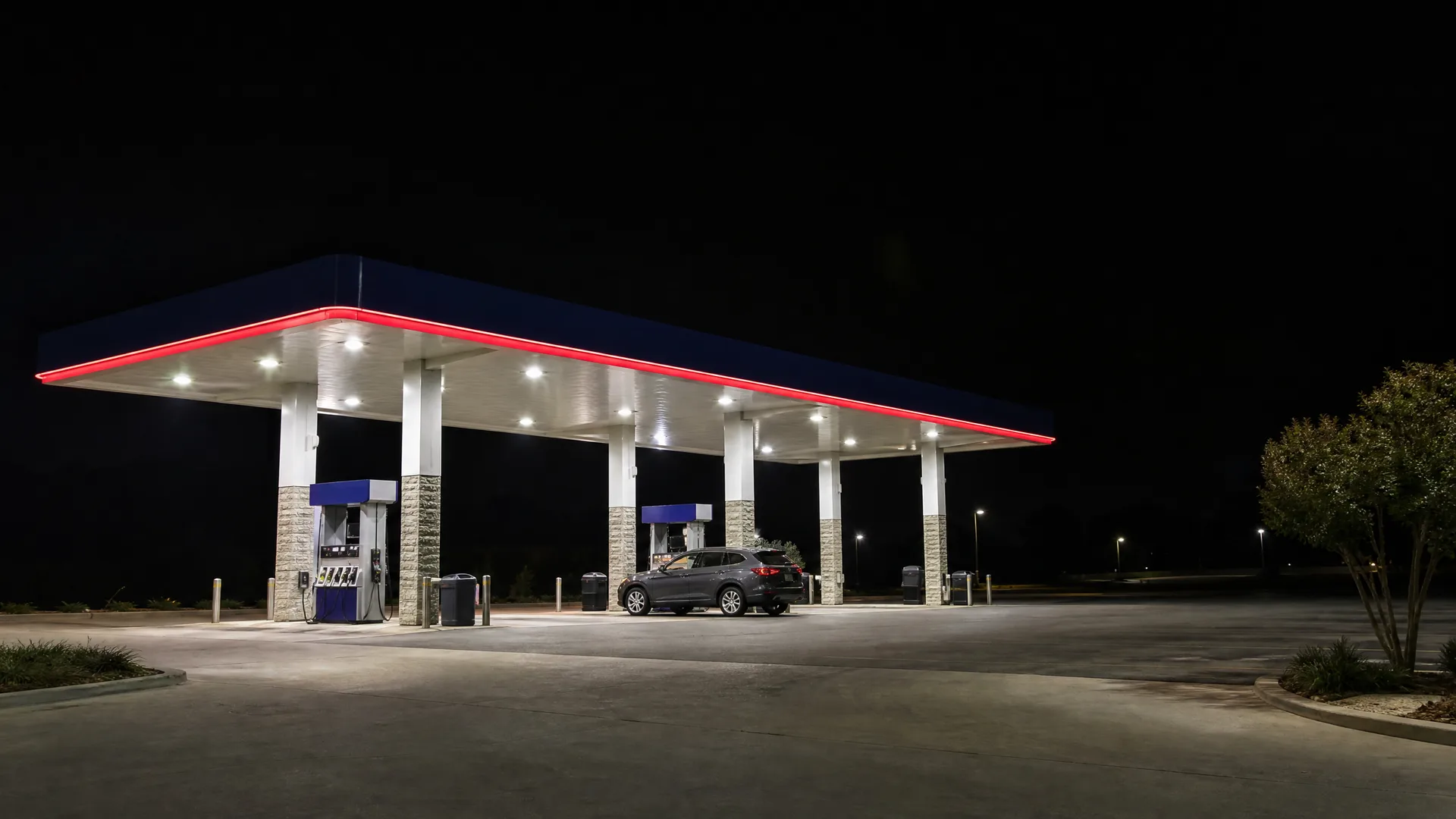 A lit gas station canopy with red trim at night, with a dark SUV parked at one of the pumps and empty paving in the foreground.