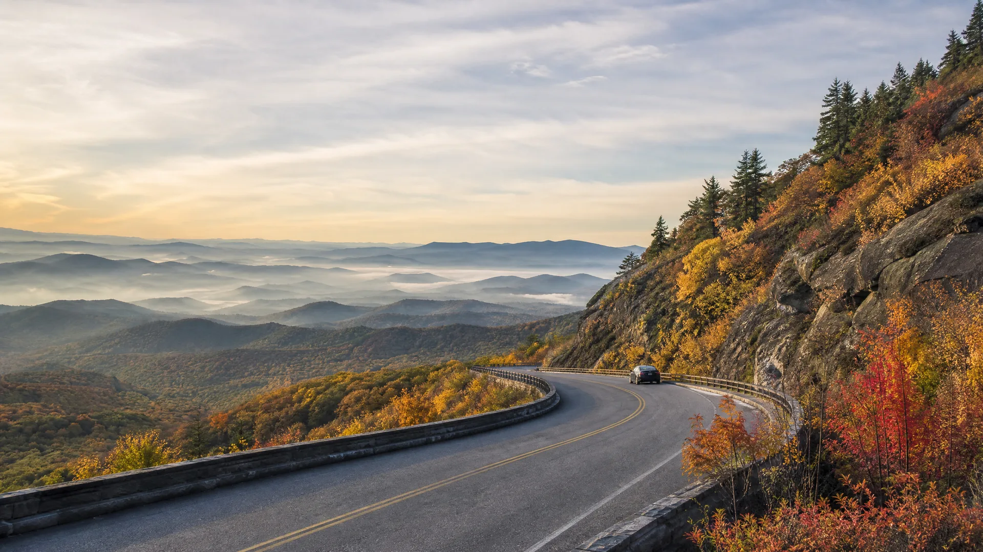 A curving two-lane mountain road with a stone guardrail runs along a rocky slope with autumn-colored trees, overlooking fog-filled valleys and layered ridges under a pale sky.