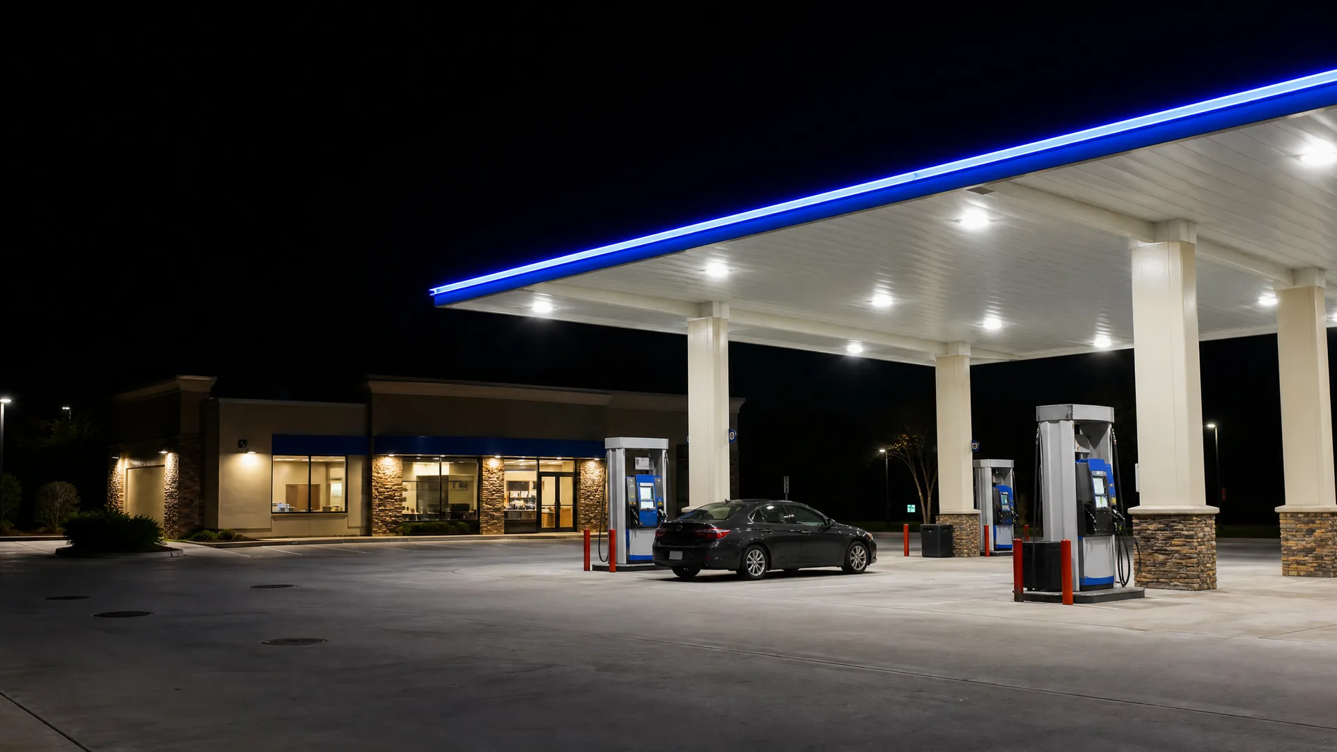A gas station at night with a lit canopy edged in blue, two fuel pump islands, a dark sedan parked near the convenience store building, and empty concrete pavement in the foreground.