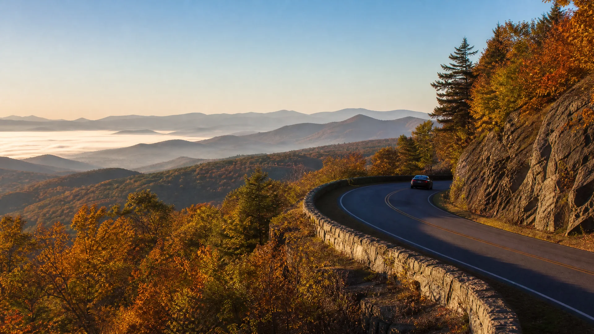 A dark car drives on a curving two-lane mountain road bordered by a stone wall and rock cliff, with autumn-colored trees and layered mist-filled valleys and ridges in the distance.