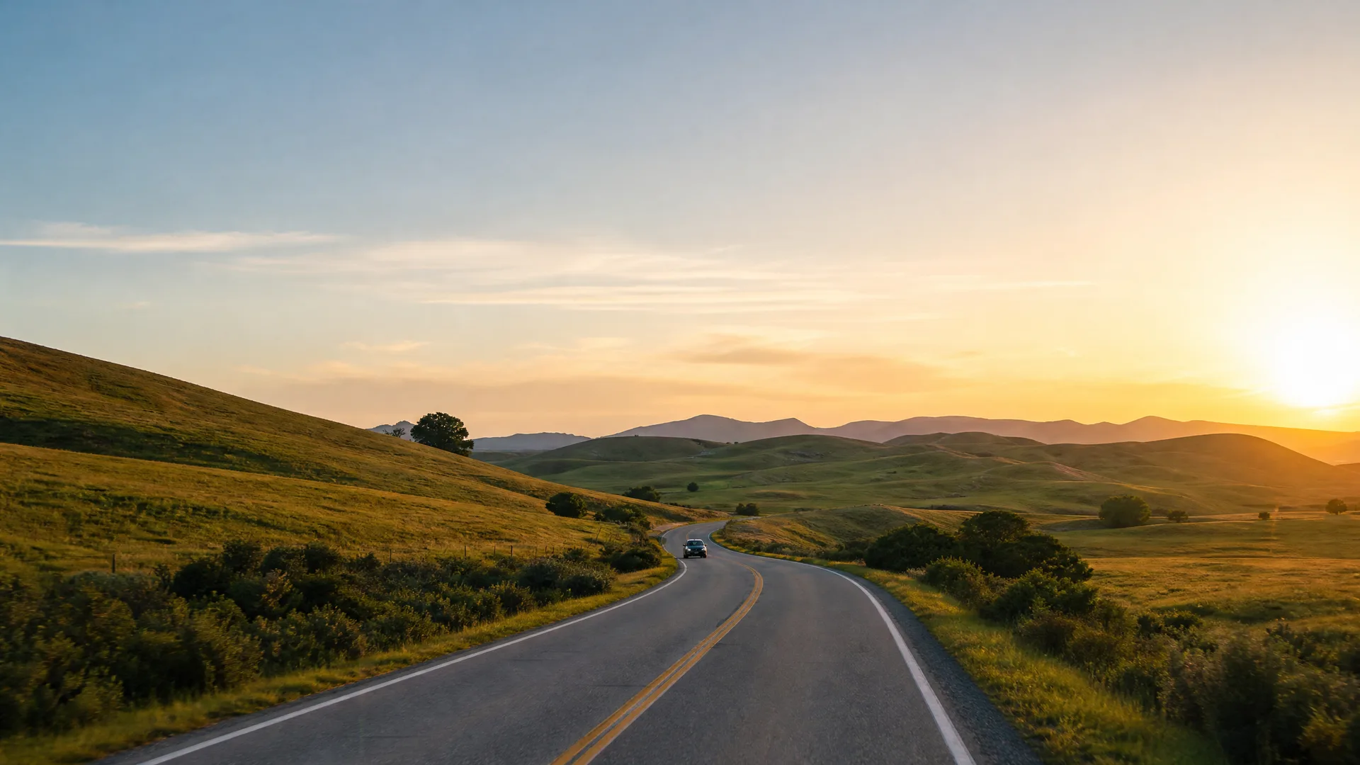 A two-lane road with a double yellow center line curves through rolling green and golden hills toward low mountains, with a single car in the distance and the sun low on the horizon.