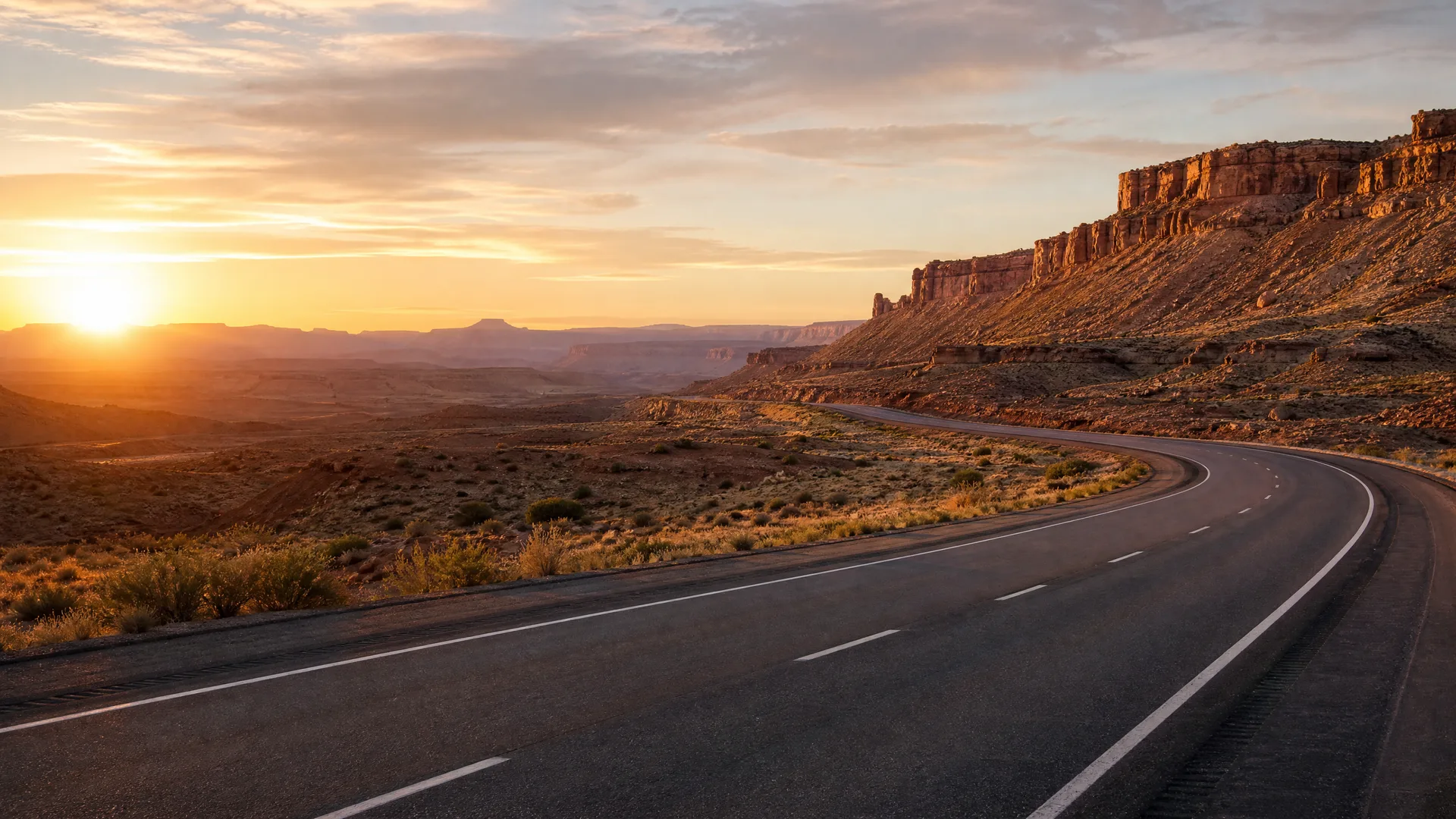An empty two-lane paved road curves through an arid desert landscape with sandstone cliffs on the right and the sun low on the horizon at left.