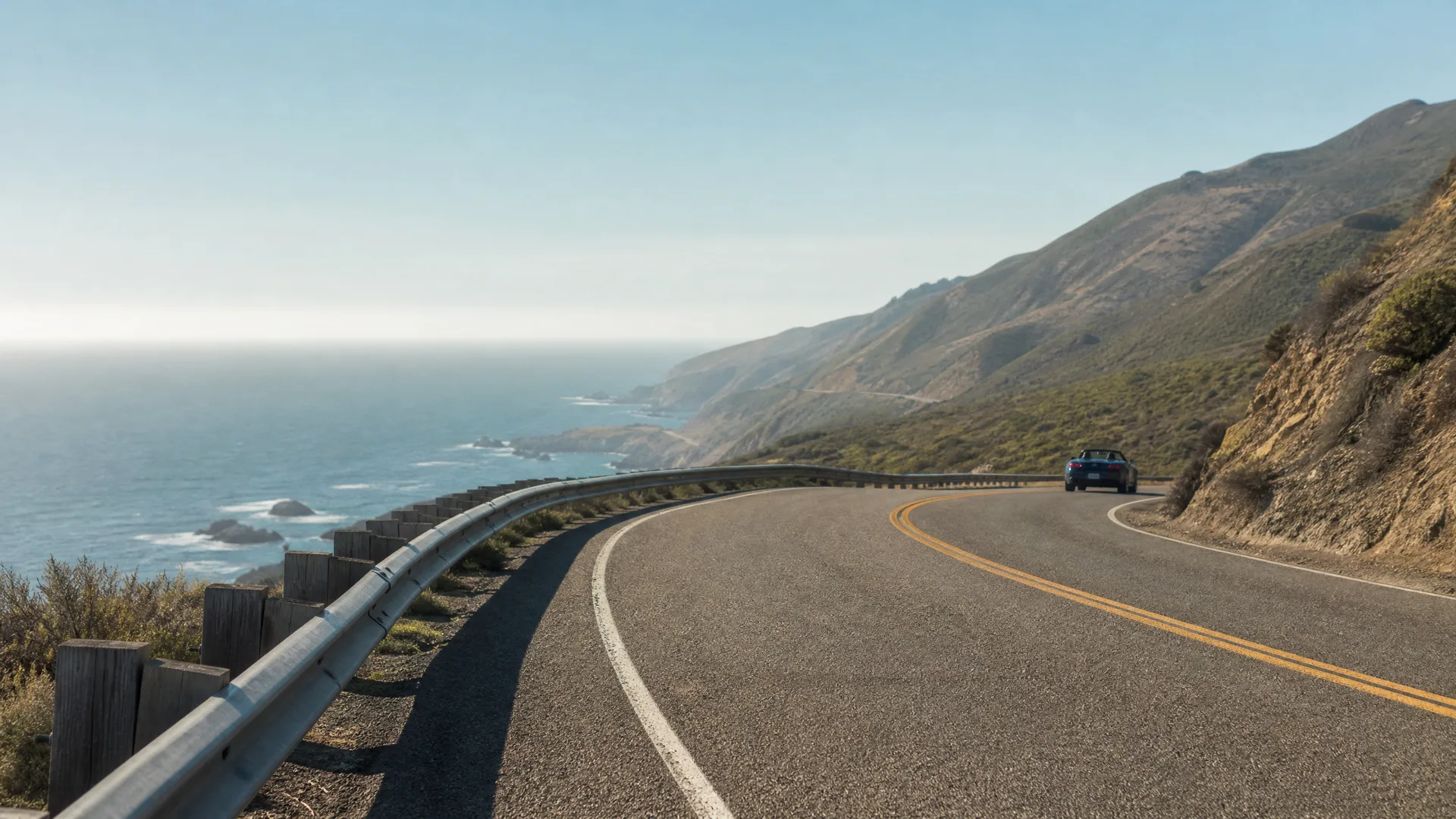 A two-lane coastal highway curves along a hillside above the ocean, with a metal guardrail and wooden posts on the left and a dark blue car driving away in the distance.