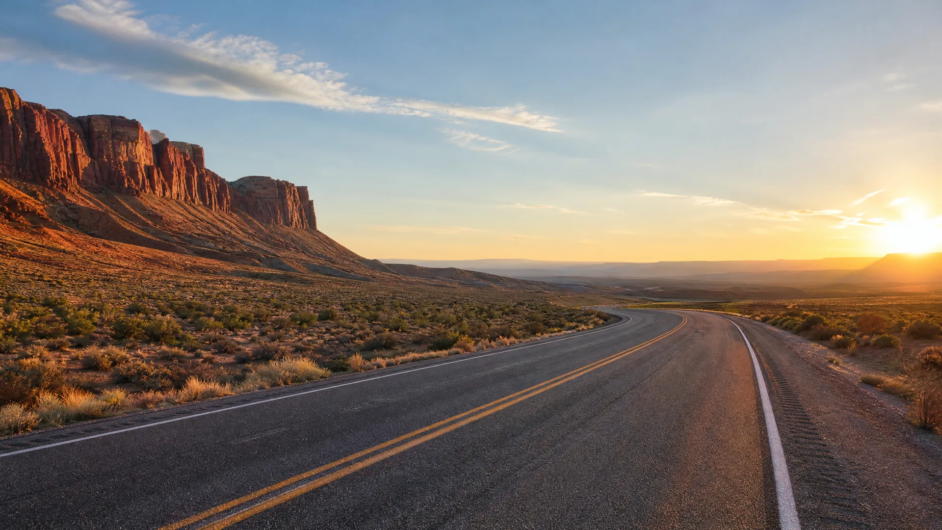 A two-lane asphalt road with double yellow center lines curves through desert scrub toward a low sun, with red sandstone cliffs rising on the left.