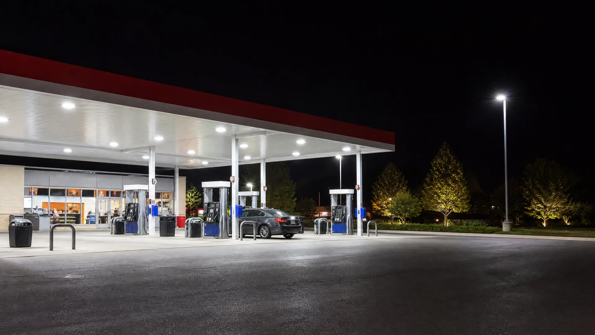 A lit gas station canopy with fuel pumps and a single dark car at night, with a convenience store at left and an empty paved lot in the foreground.