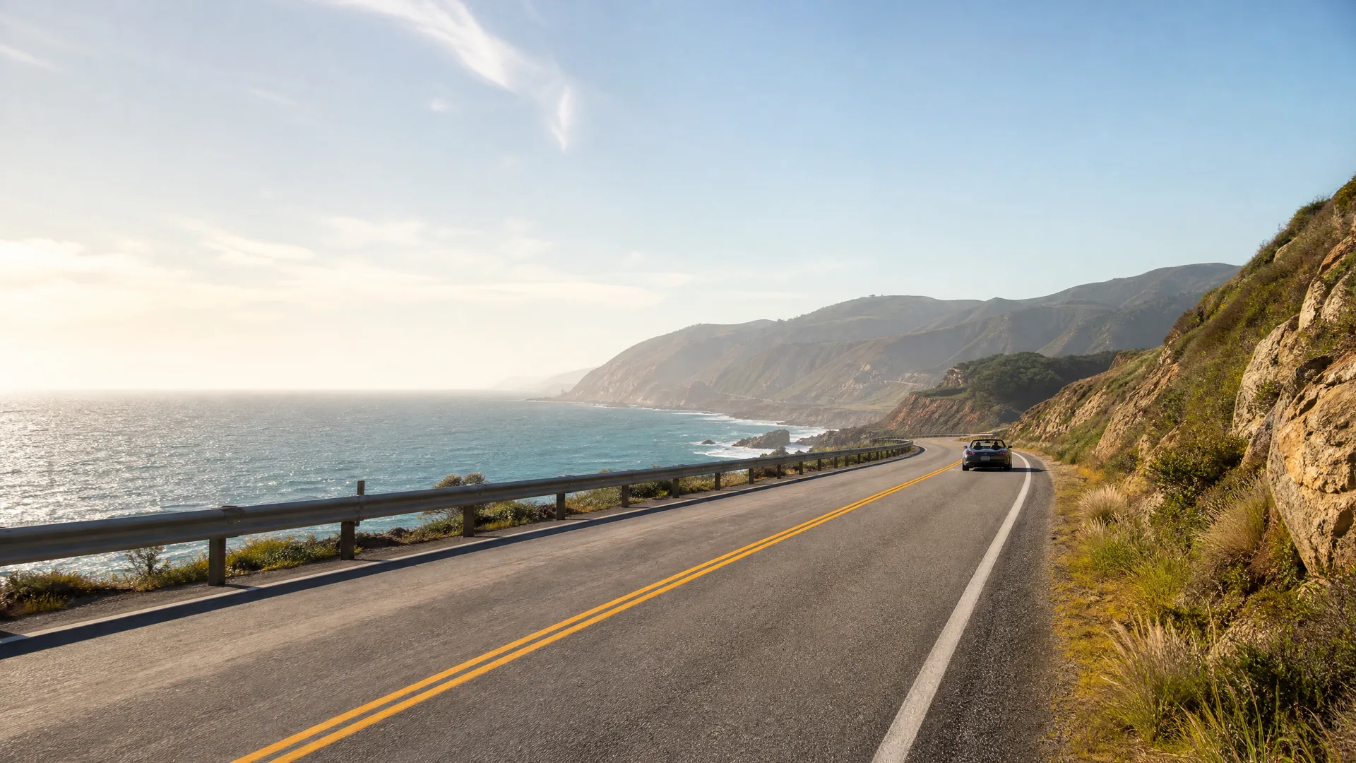 A two-lane coastal highway with double yellow center lines curves along a hillside above the ocean, with a dark convertible car driving away in the distance and hills along the shoreline.