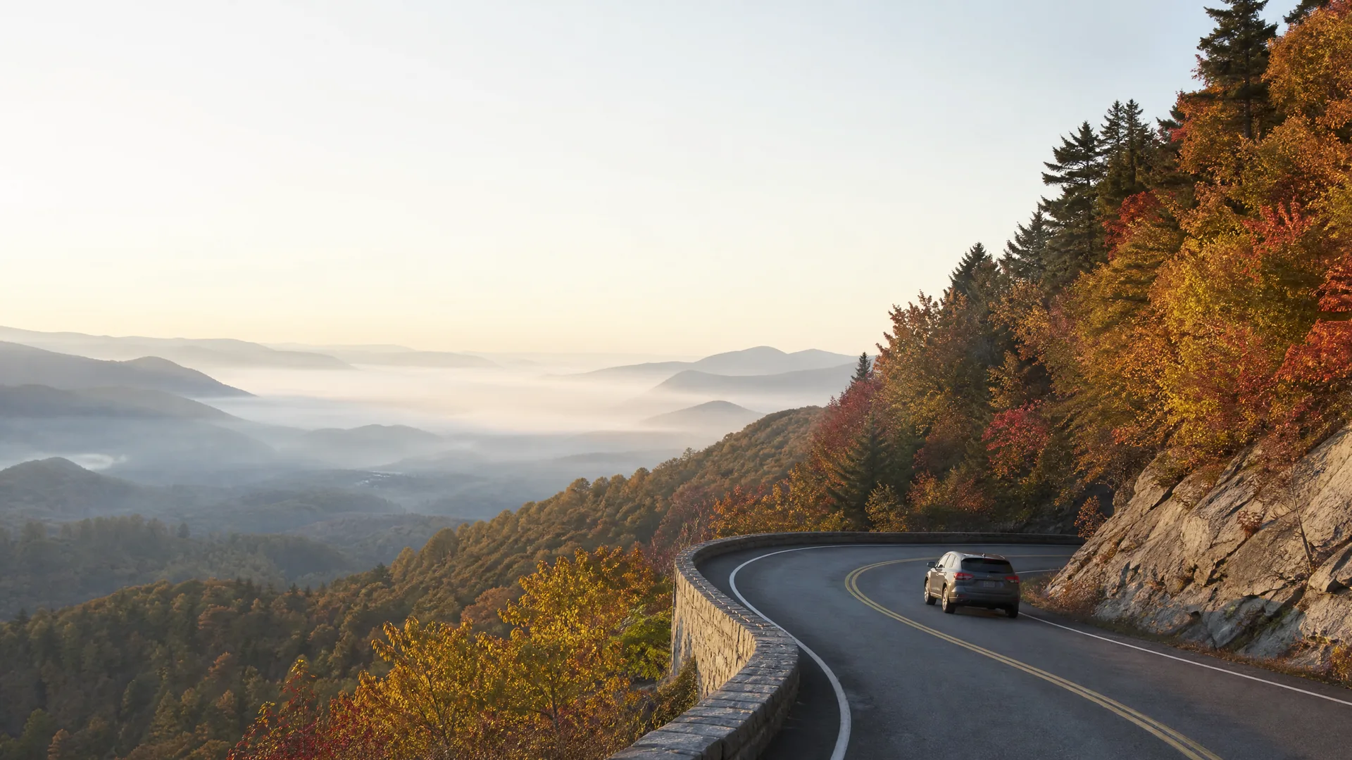 An SUV drives on a curving two-lane mountain road with a stone retaining wall, bordered by autumn-colored trees and a rock face, overlooking fog-filled valleys and distant ridges at sunrise.