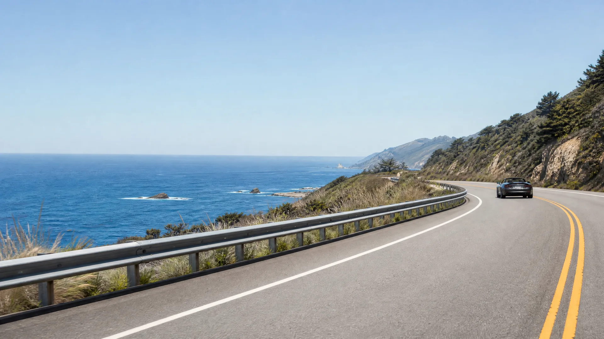 A dark convertible car drives along a two-lane coastal highway with a metal guardrail, with the ocean and rocky, tree-covered hillsides in the background under a clear sky.