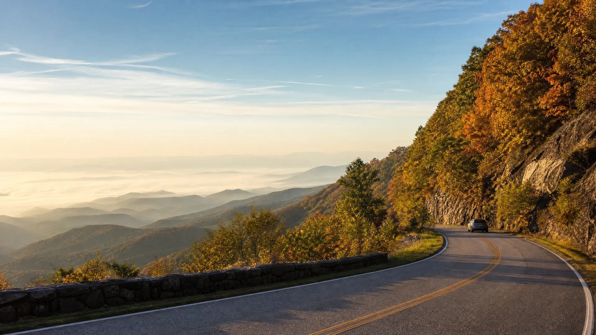 A two-lane mountain road with a double yellow line curves along a wooded ridge with autumn-colored trees and a low stone wall, overlooking layered hazy hills and valleys under a clear sky, with a single dark car in the distance.